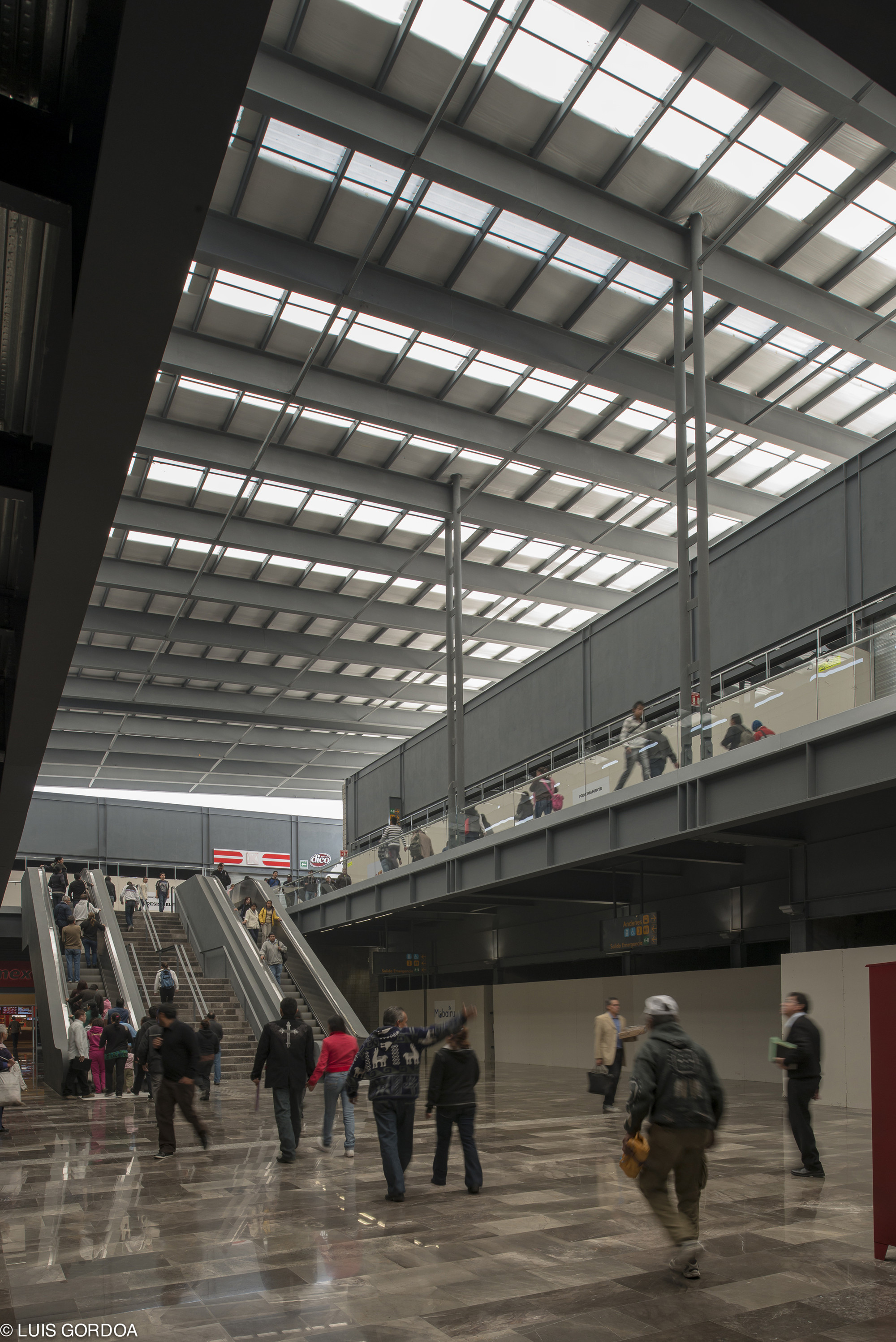 Double-height hall with skylights overhead, escalators on the left and a mezzanine bridge, as travelers cross the polished stone floor