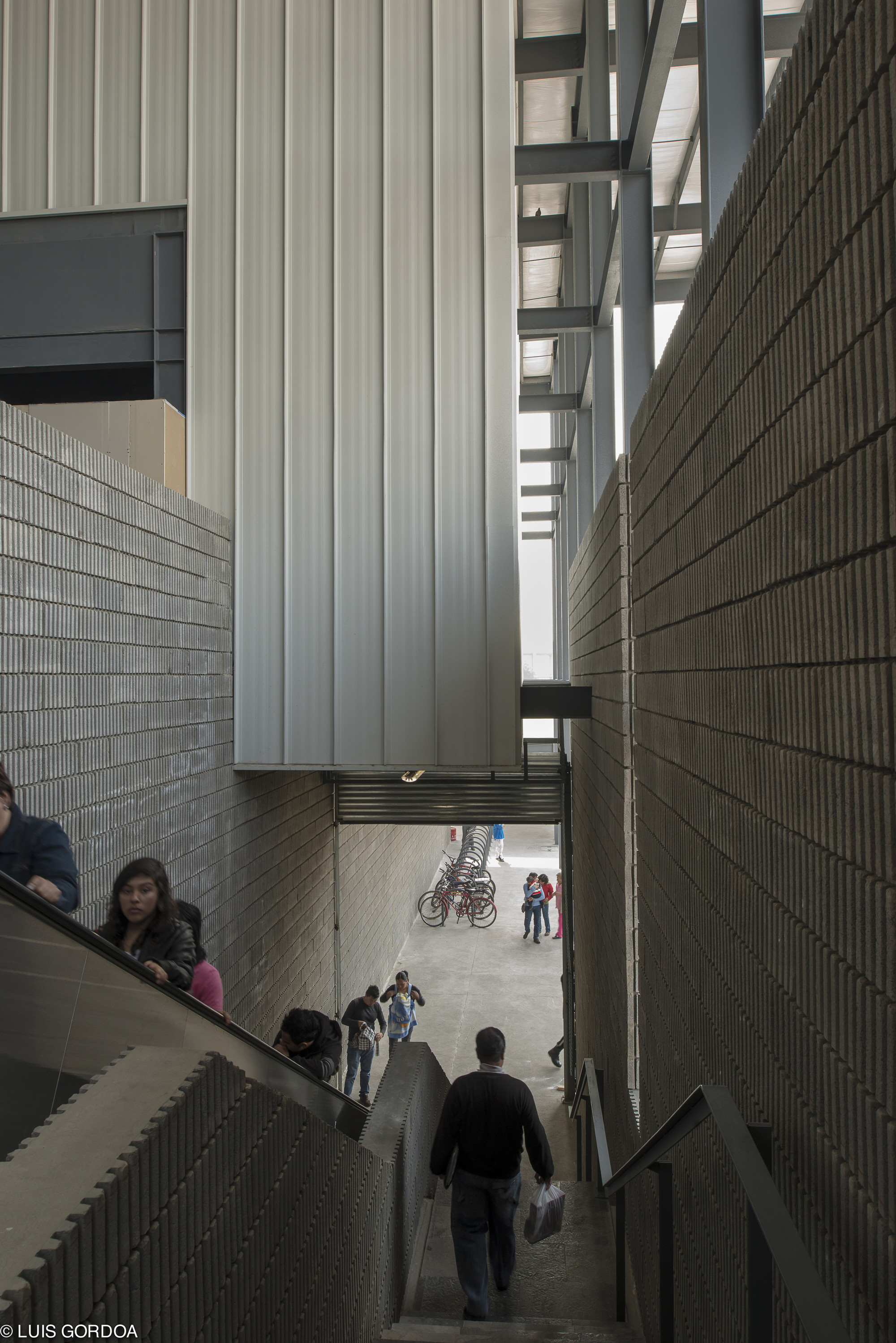 Narrow stairway between ribbed concrete block walls descending toward a bicycle rack, with a pedestrian carrying a bag in daylight
