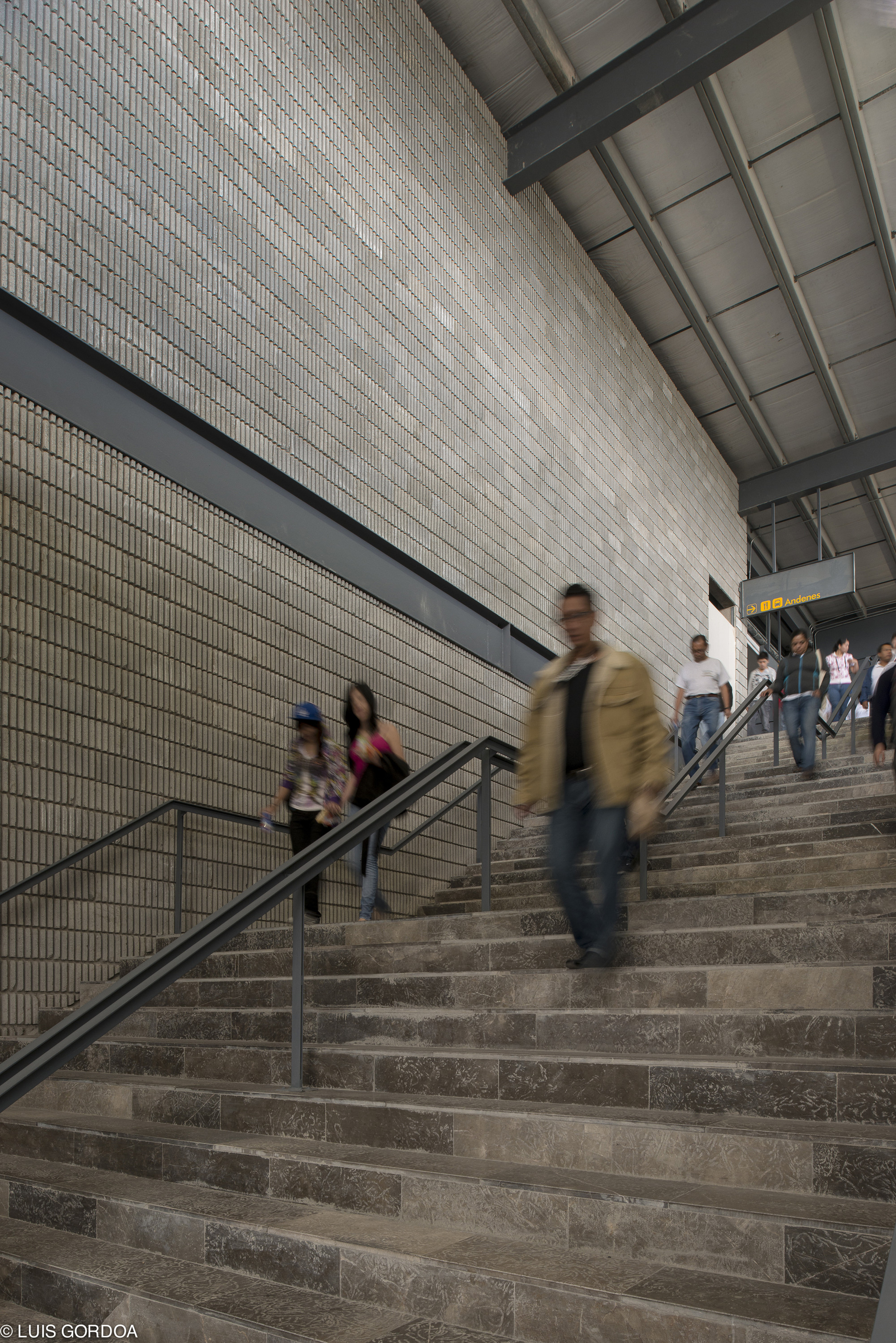 Commuters descending a wide stone staircase beside a tiled brick wall and steel handrails under a sloped ceiling, one figure blurred mid-step