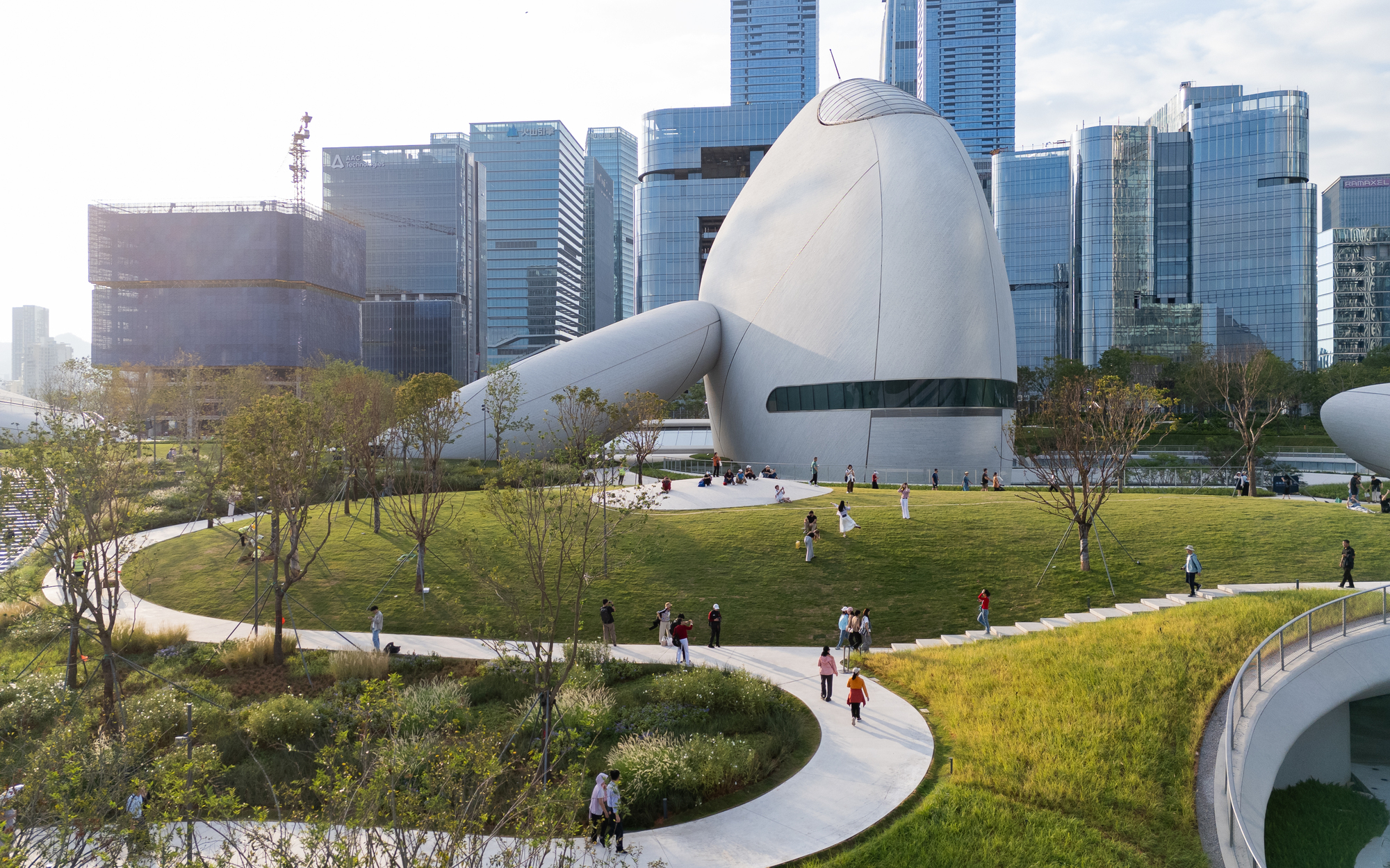 Egg-shaped white volume with a ribbon window rising from a grass terrace, with visitors walking a curving path before glass towers