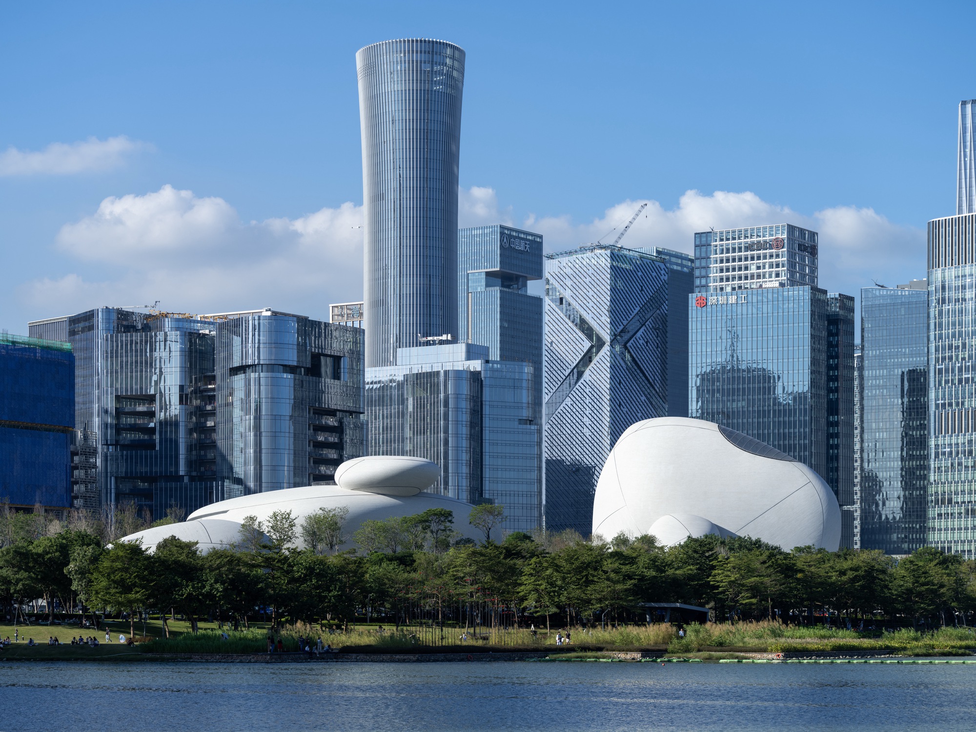 Two white pebble-shaped roof forms sit below a cluster of glass towers, seen across a lake under blue sky
