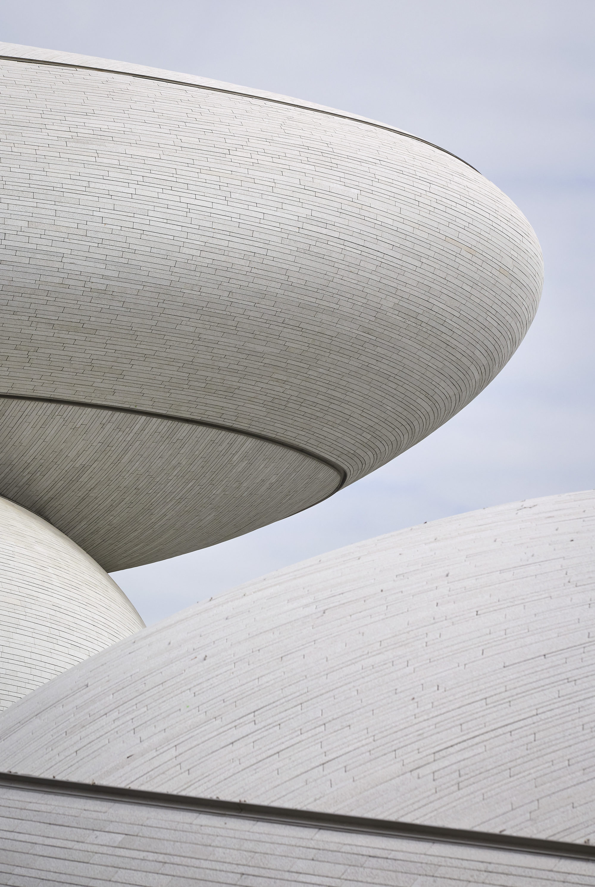 Close-up of overlapping white domes clad in thin stone strips, with a saucer-shaped overhang against a pale cloudy sky