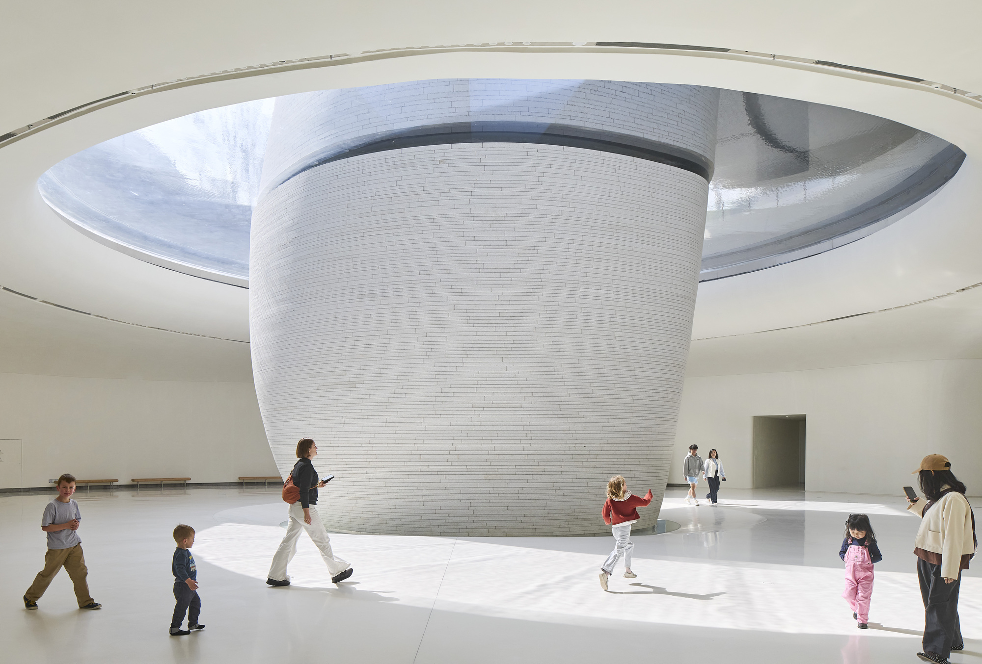 Interior with a cylindrical white stone-clad core rising through an oval skylight, children and adults crossing the sunlit floor
