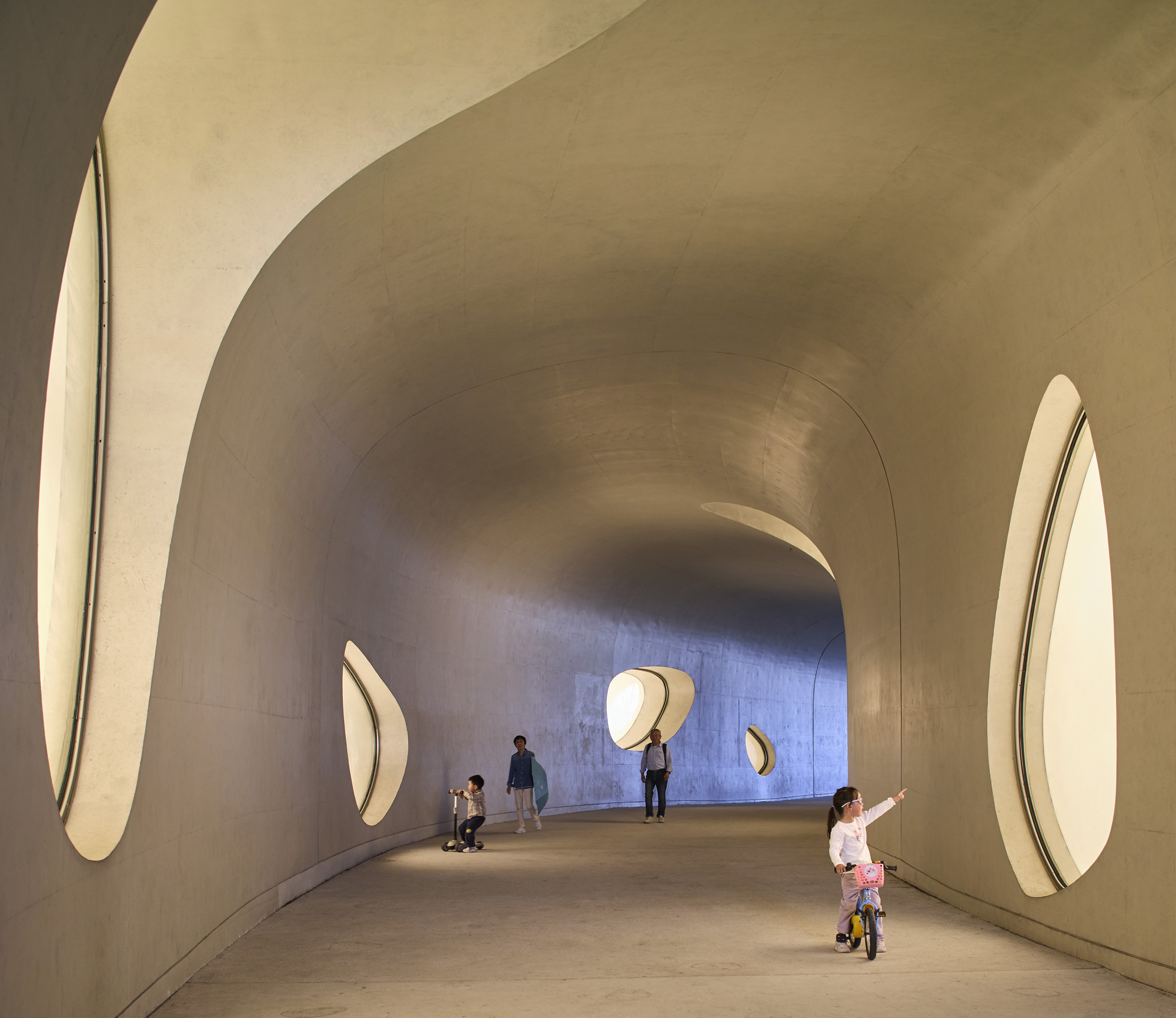 Curving concrete passage with oval window openings, a child on a balance bike pointing and others walking toward the far end