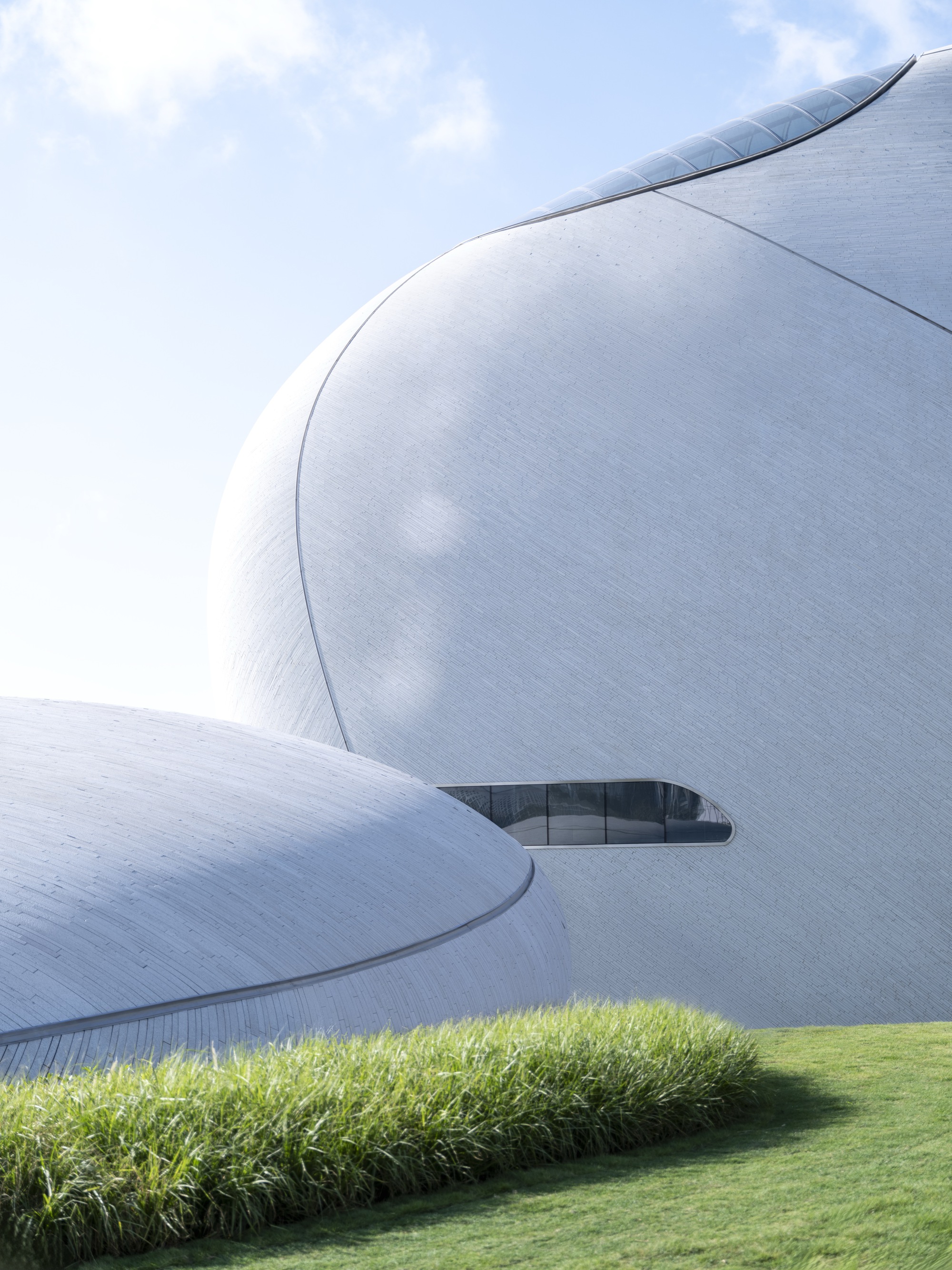 Detail of two white stone-clad curved volumes with a slim ribbon window, a clipped grass bed in the foreground in bright sun