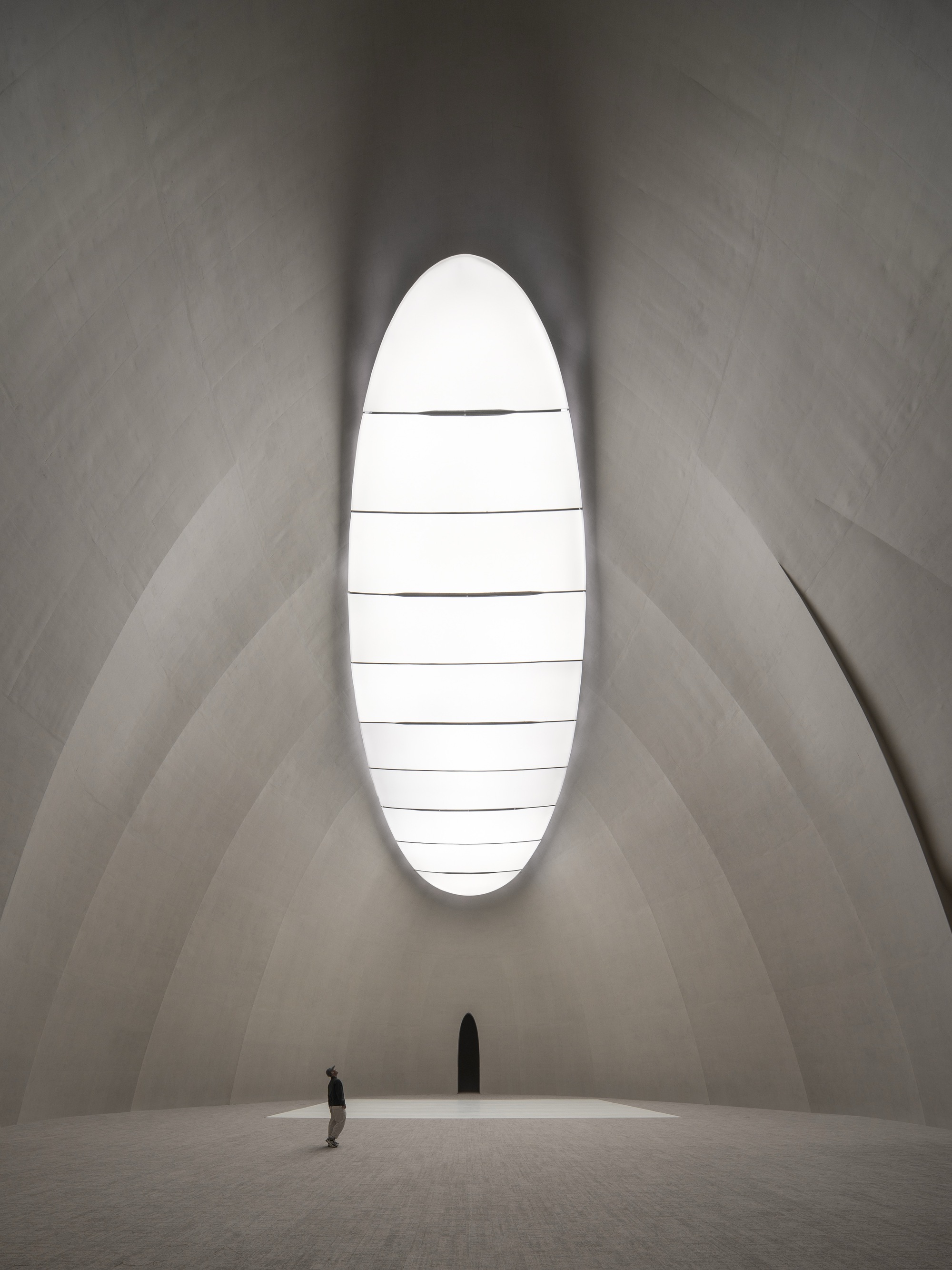 Tall oval skylight in a pale curved concrete vault, with a lone visitor looking up from the floor below