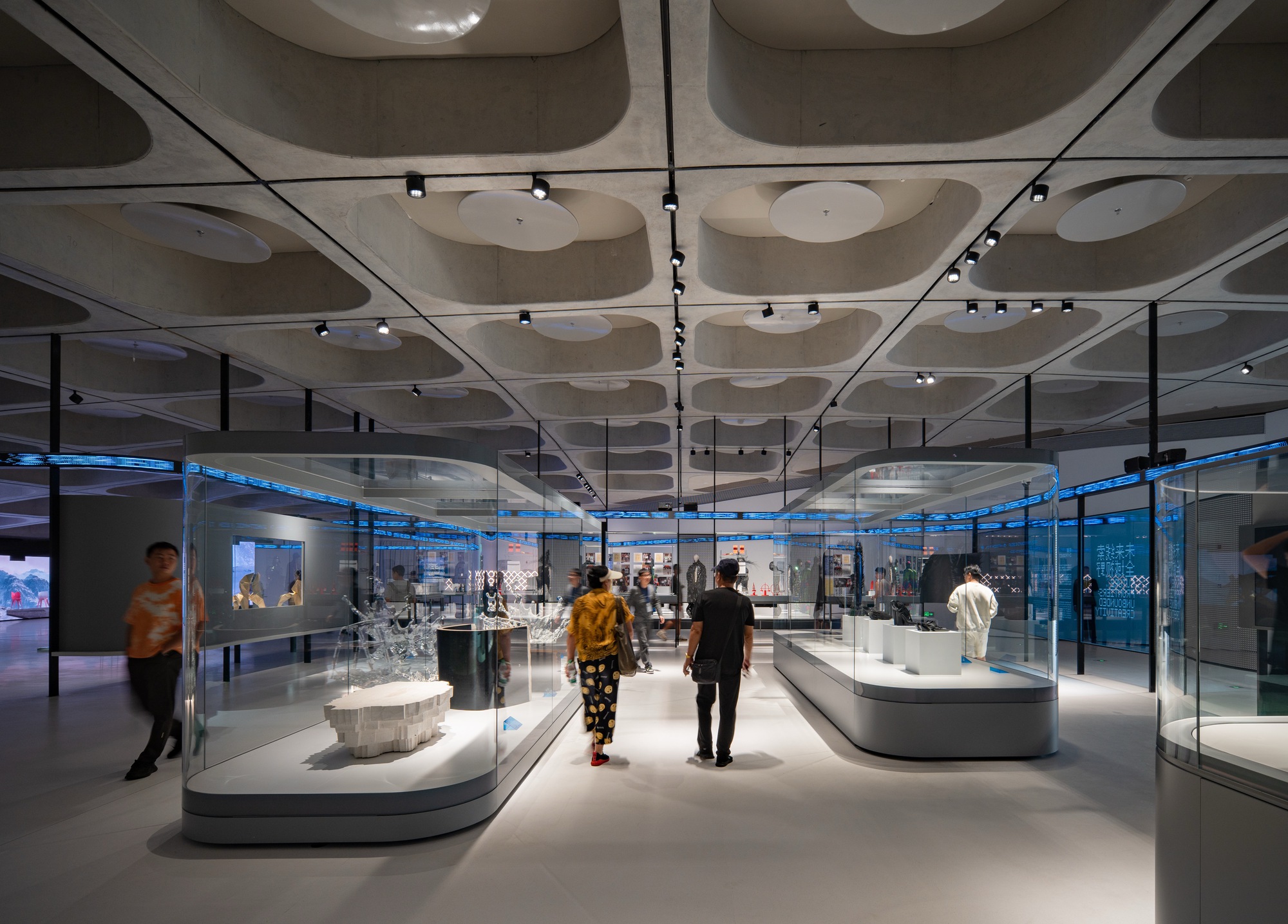 Exhibition hall with curved glass display cases under a concrete ceiling of scooped coffers, visitors walking between the vitrines