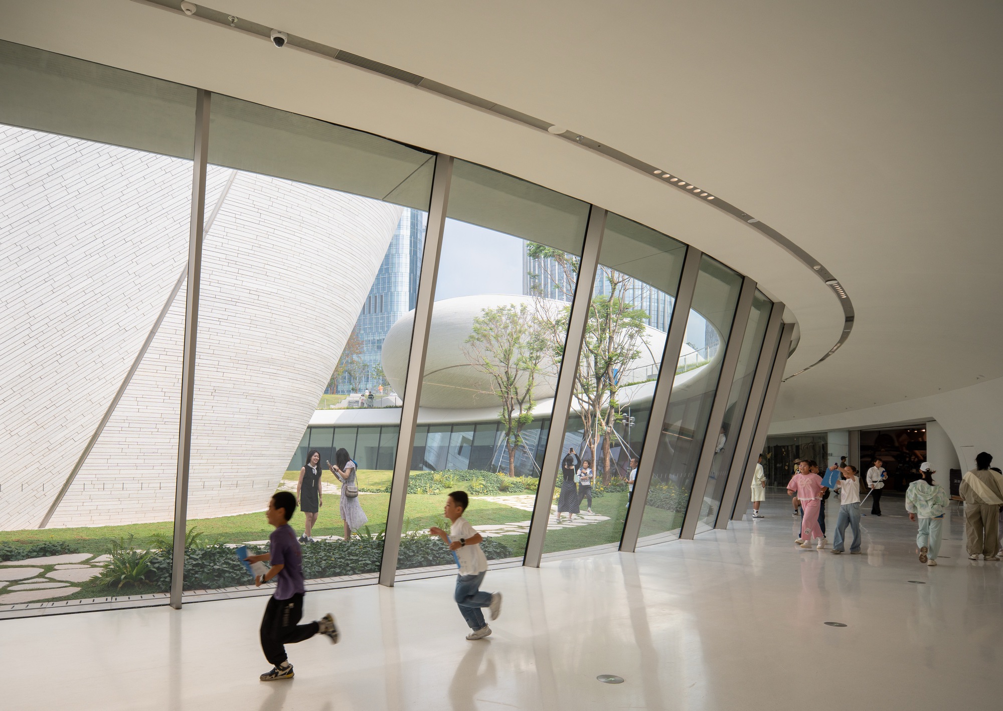 Curved glazed wall of a bright concourse looking onto a planted garden and white tiled forms, children running past