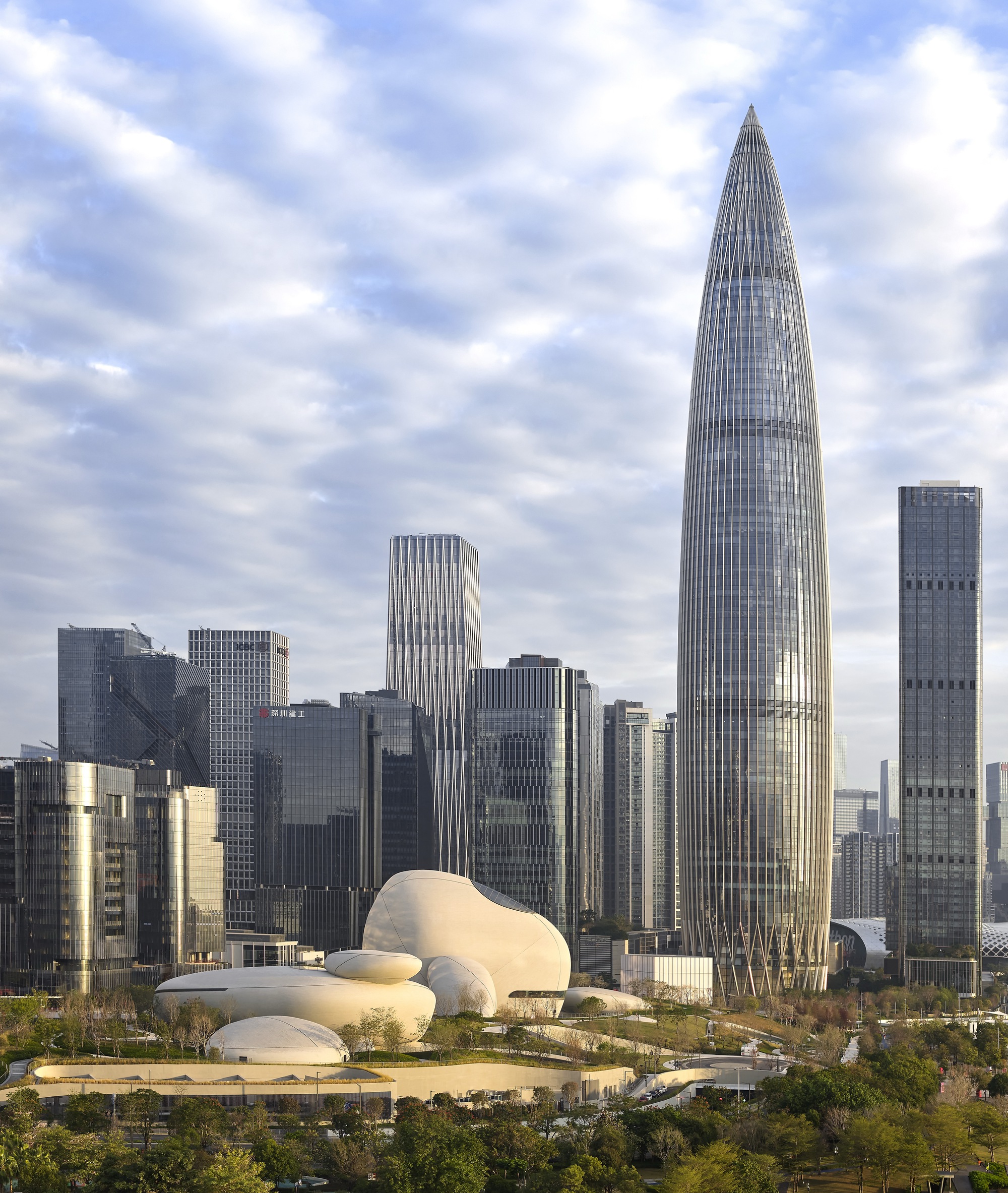 Skyline view of a tapering glass tower beside low white rounded volumes among park trees under a cloudy sky