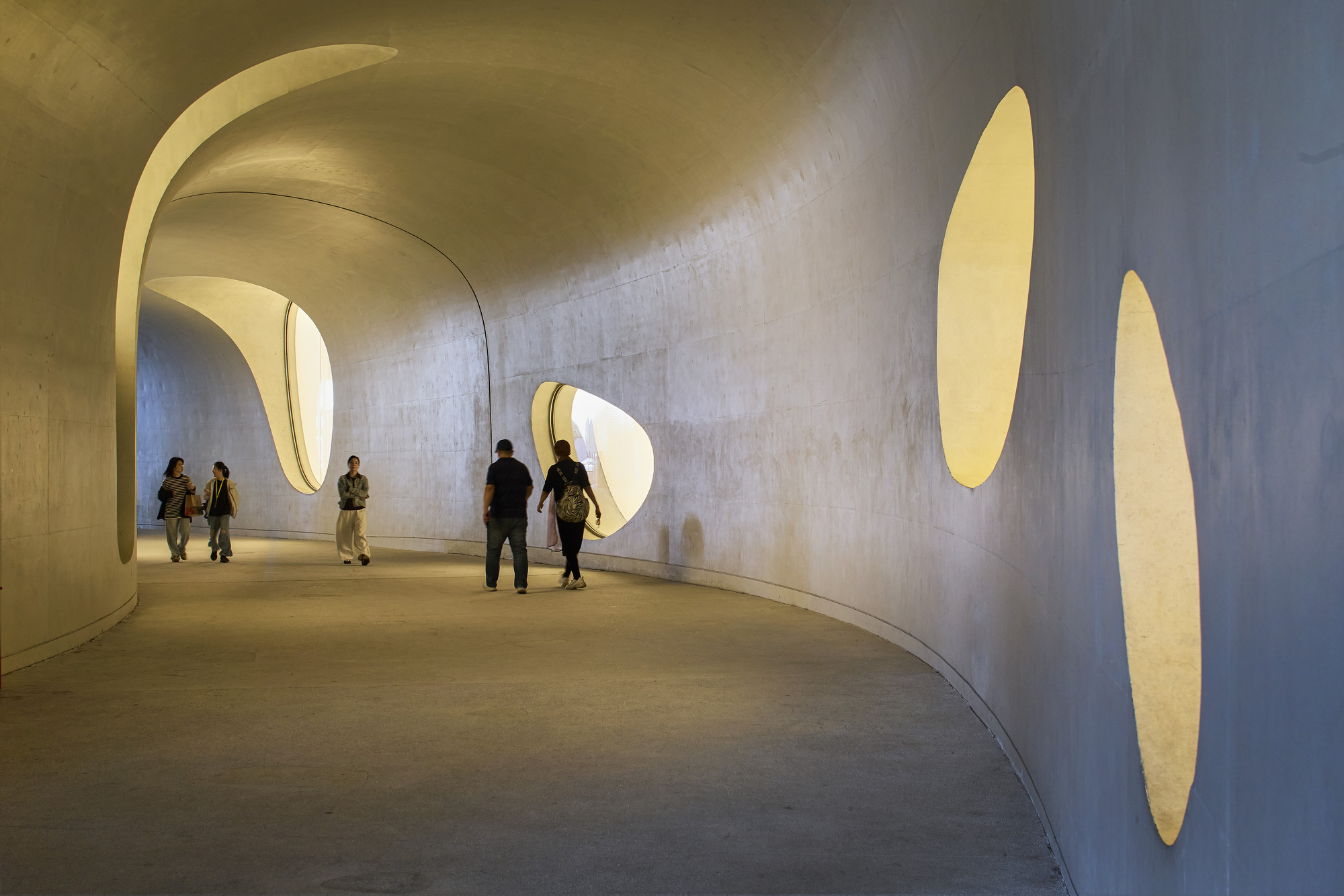 Curving concrete passage with oval openings glowing yellow in the walls, a few people walking through in warm light