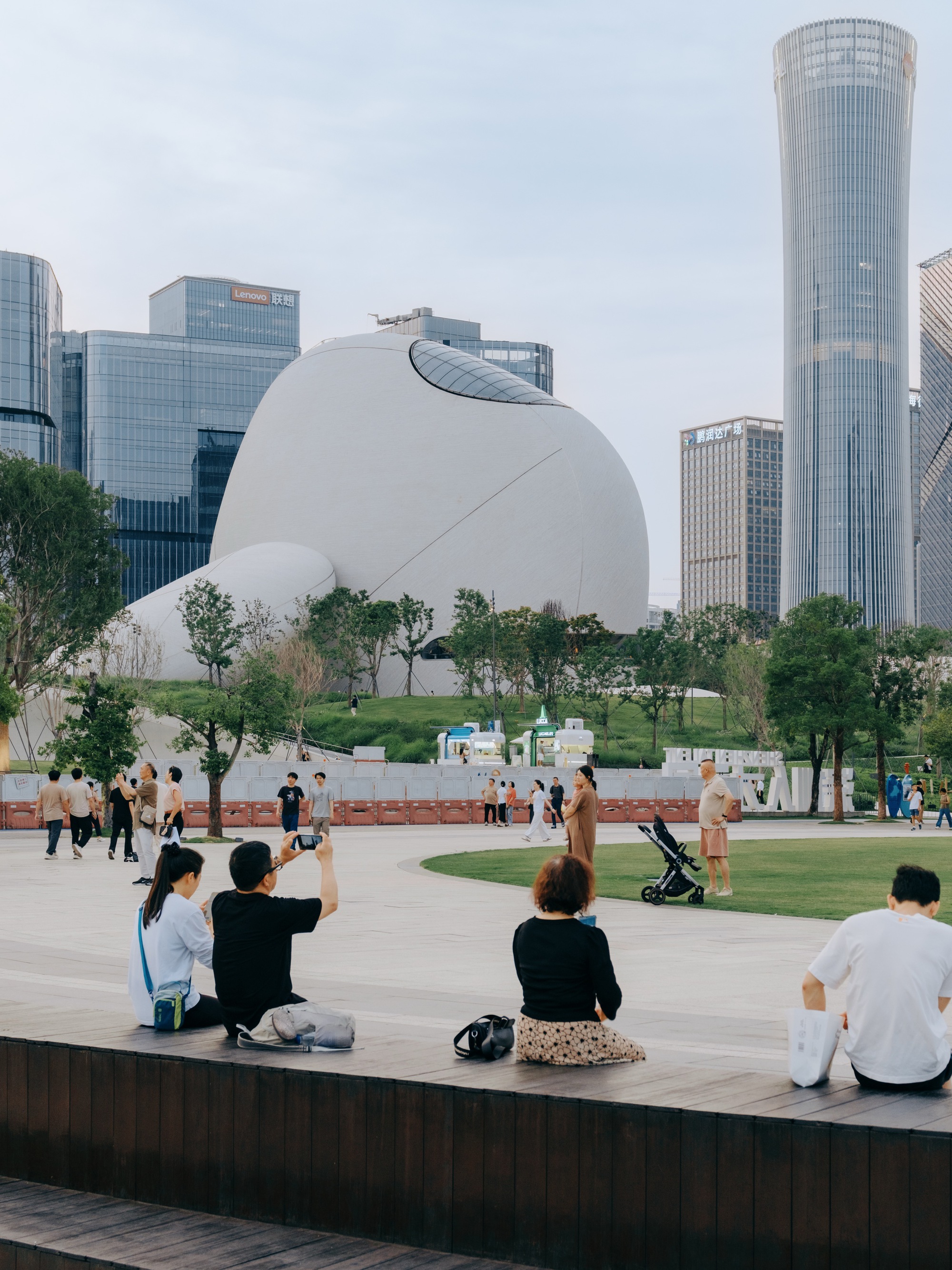 Visitors sitting on a timber bench in a plaza facing a white domed building with a skylight, towers rising behind
