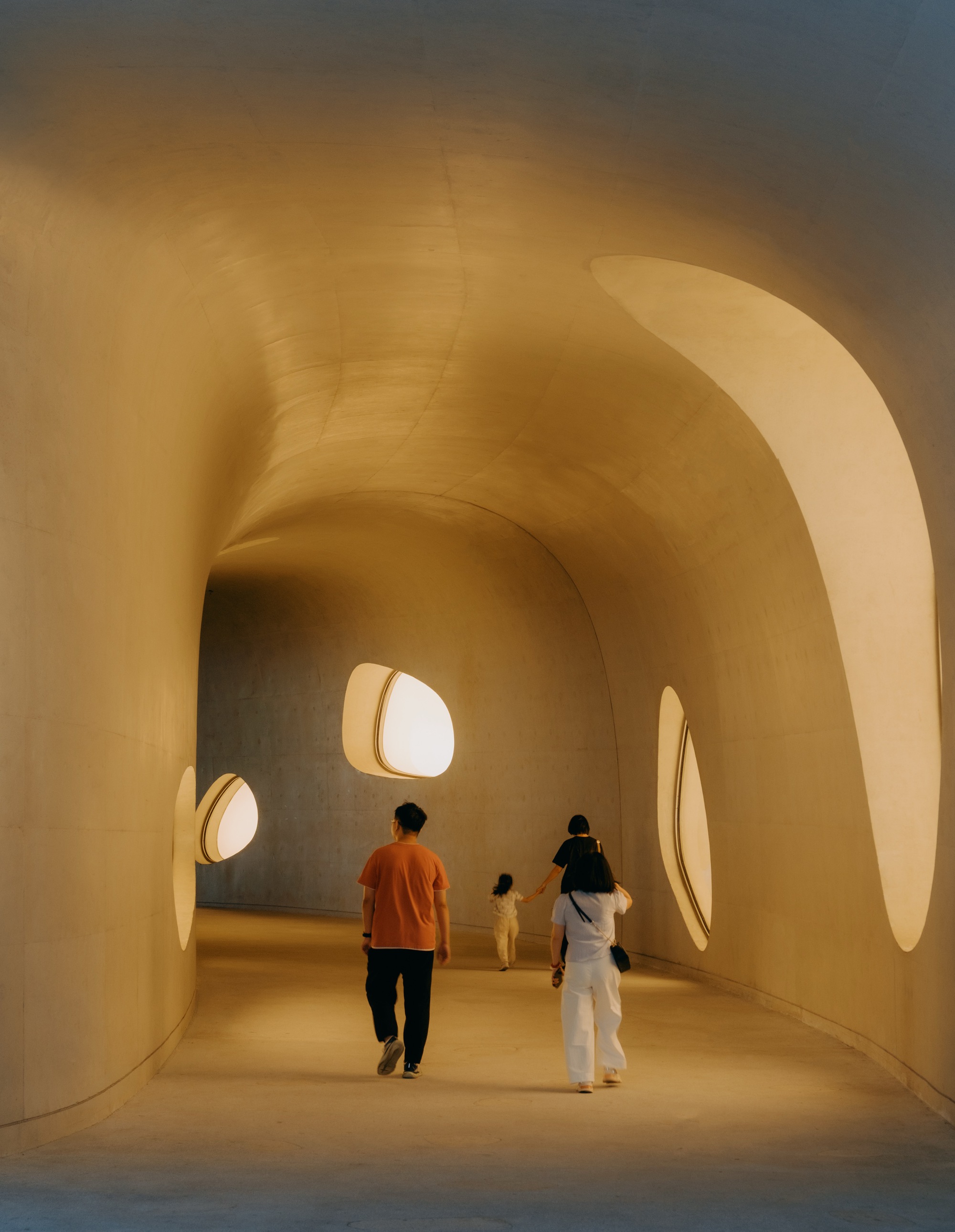 Curving concrete corridor with oval skylit openings as a family walks toward the far end in warm evening light