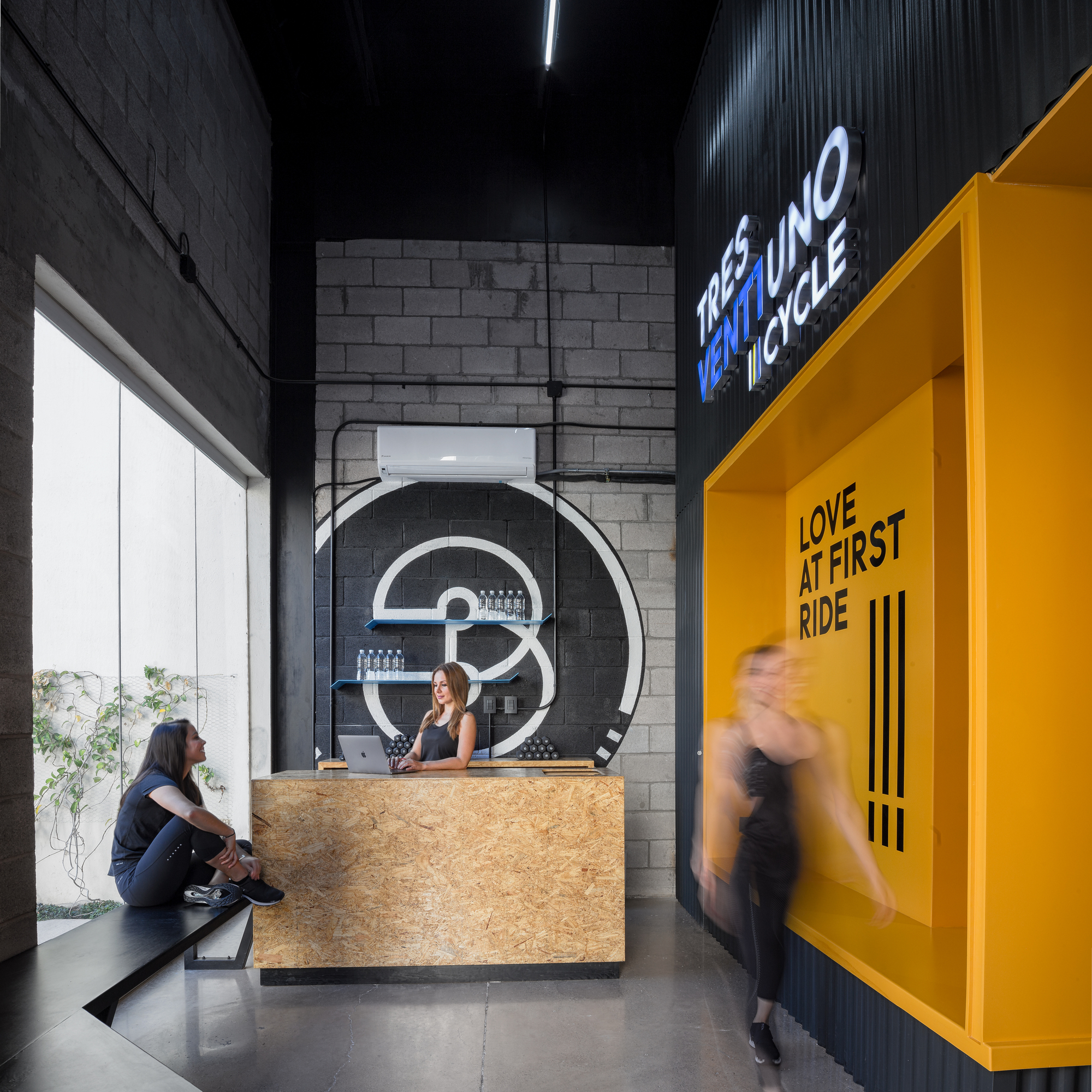 Reception desk of OSB panels beneath a painted logo on a block wall, with a blurred passerby beside a yellow alcove