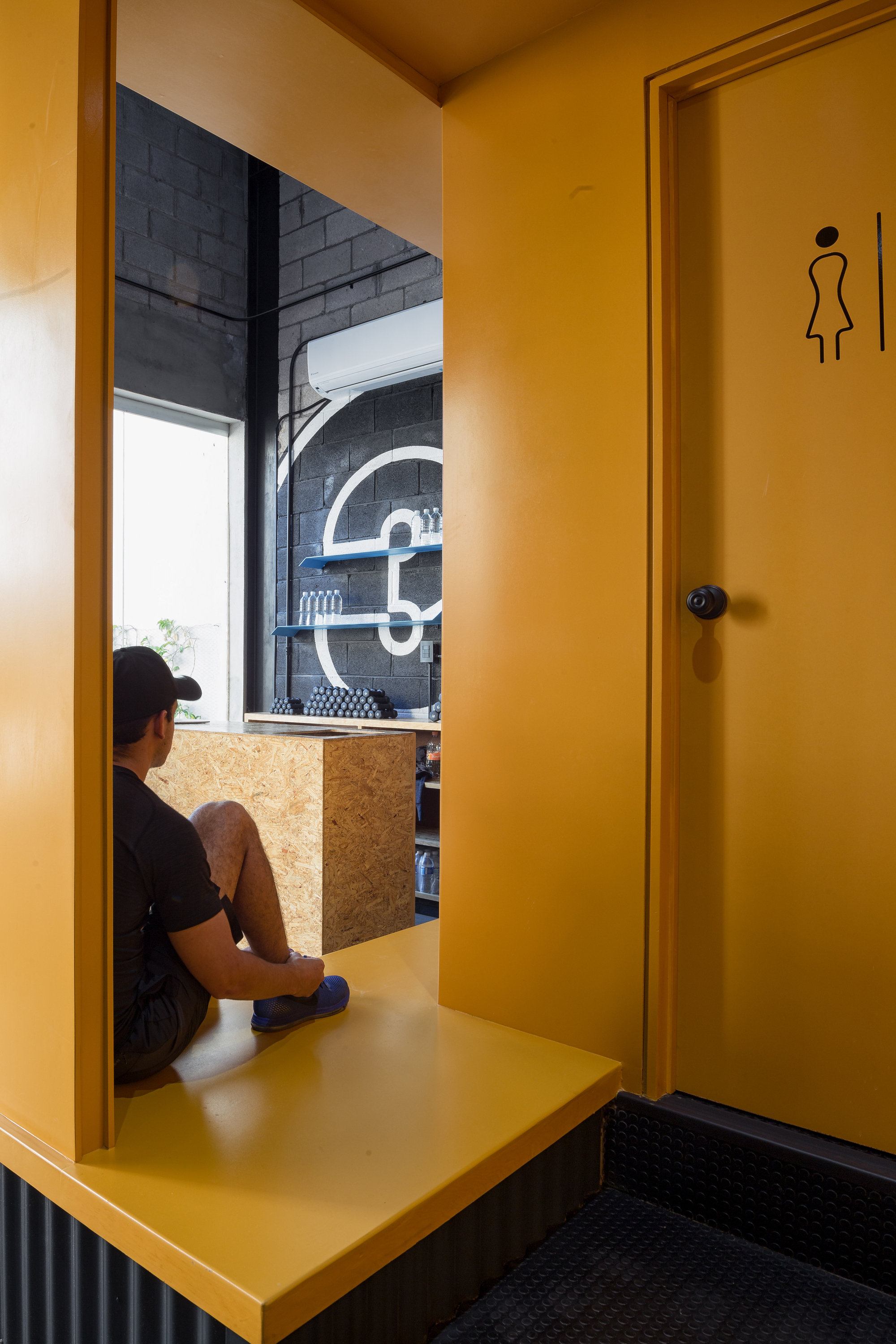Man seated on a yellow ledge looking toward the OSB counter and painted logo, beside a yellow restroom door