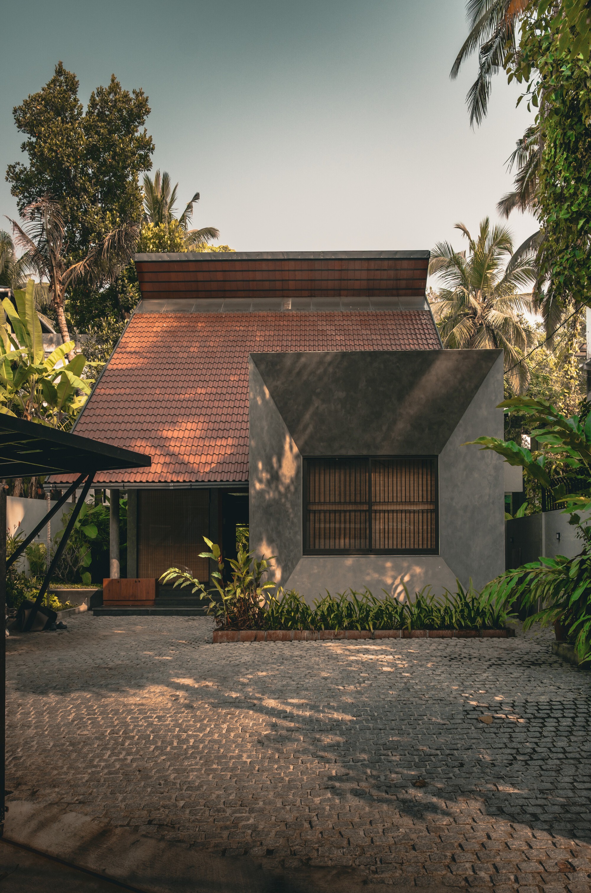 Front facade with a clay-tiled pitched roof and a grey plastered angular bay, seen across a cobbled courtyard in dappled afternoon light
