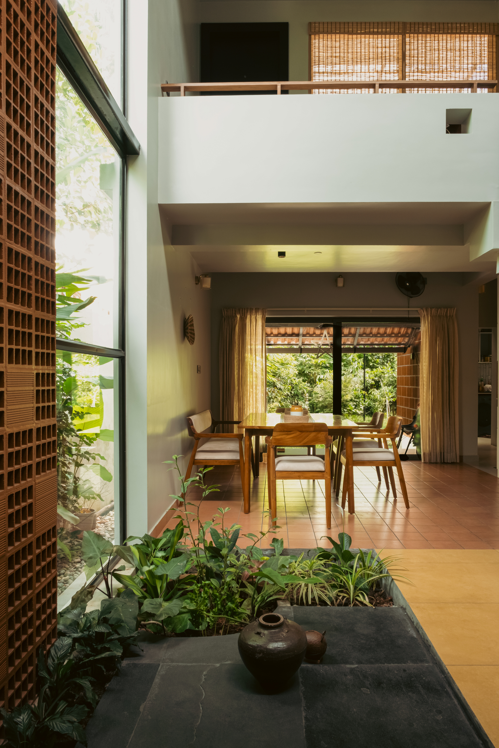 Double-height living space with a perforated terracotta screen, slate planter with a clay pot, and a dining table beyond, in soft daylight