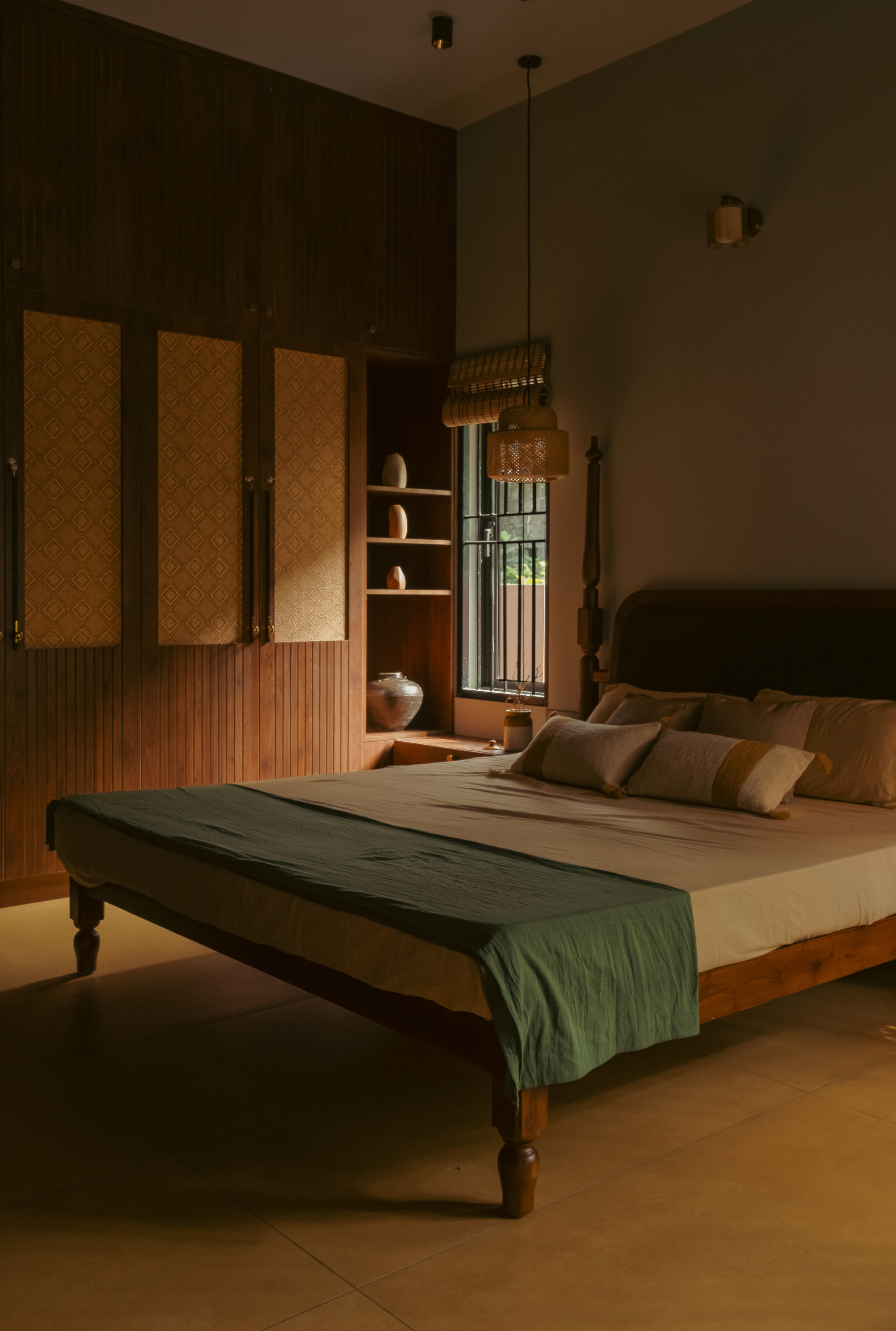 Bedroom with a low timber bed, green throw, patterned wardrobe panels and a woven pendant lamp in dim evening light