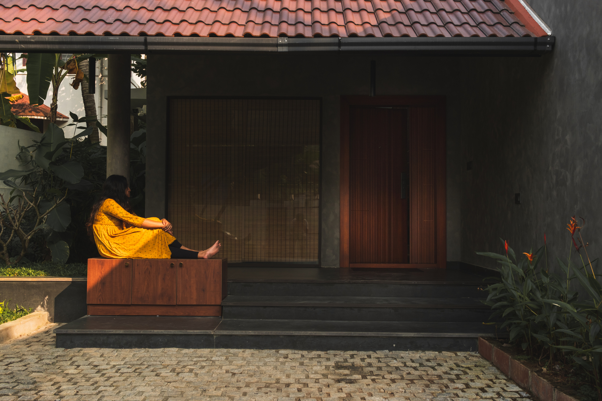 Entrance porch with a woman in a yellow dress seated on a timber bench beside a red wooden door, in warm low sun