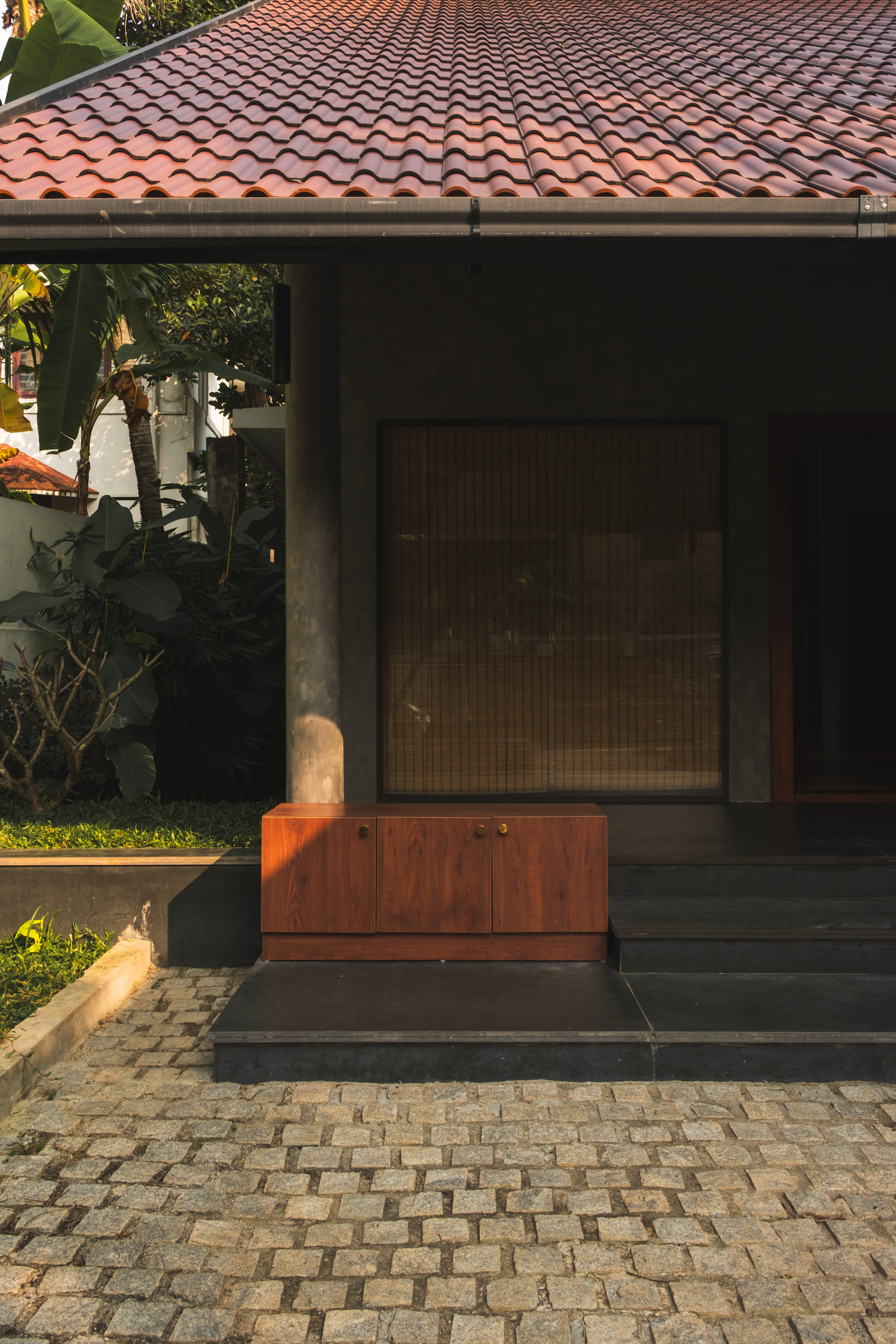 Timber storage bench on a dark stone step below a clay tile roof, with a bamboo screen and cobbled forecourt in afternoon light
