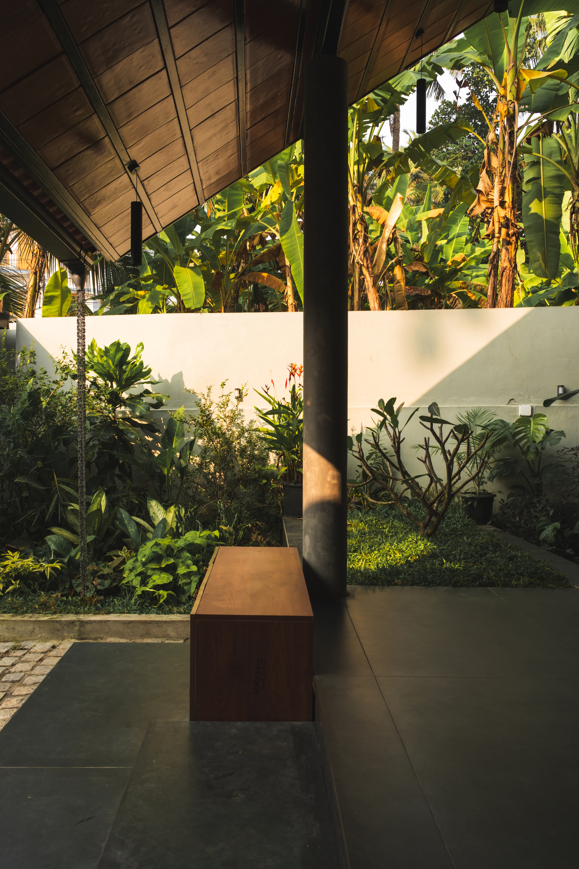 Covered porch with a concrete column and timber bench facing a garden of banana plants and frangipani in late sun