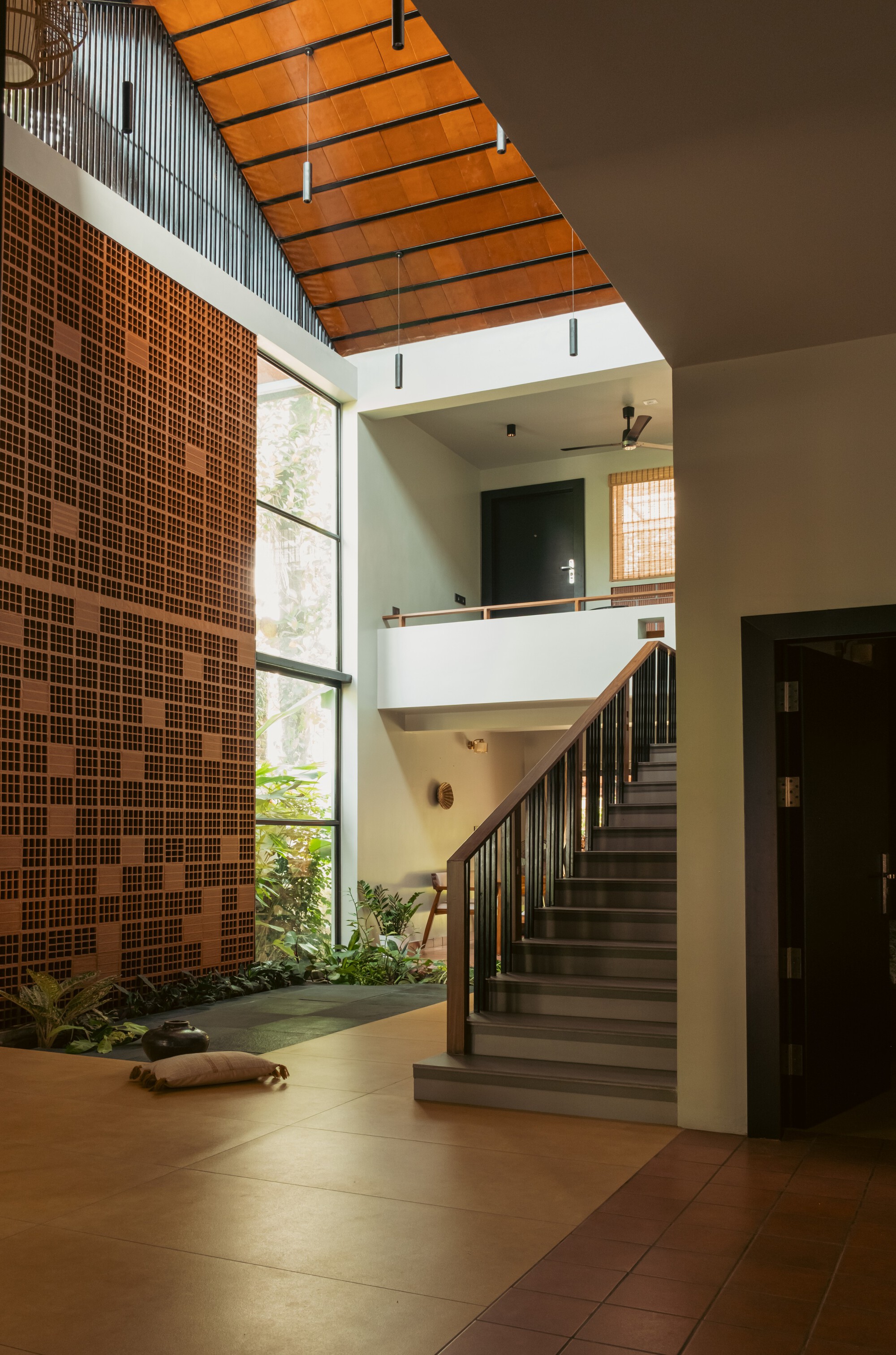 Double-height hall with a perforated brick screen wall, a timber-railed staircase and a wooden ceiling lit by daylight from a tall window