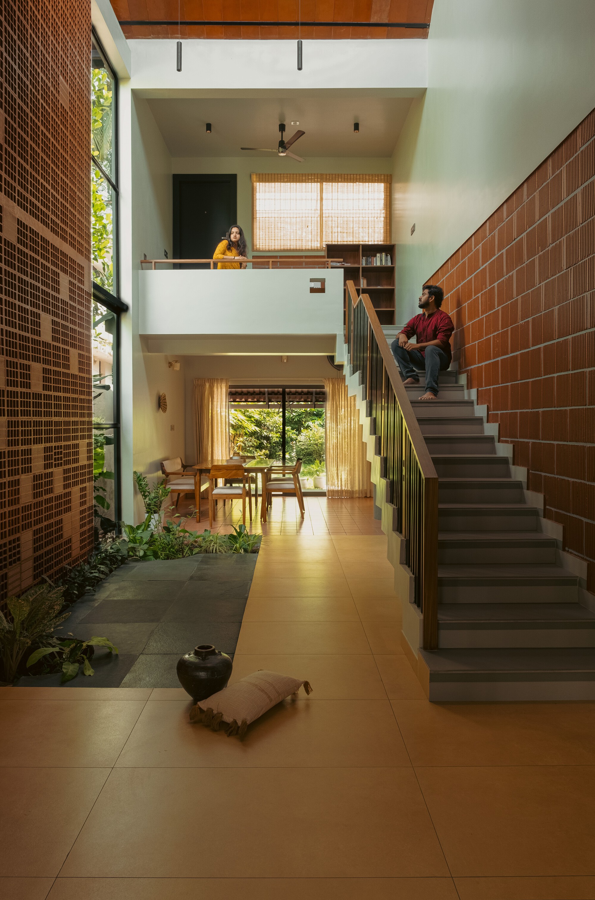 Stair hall with a man seated on the steps, a woman at the upper landing and a planted slate strip below a brick wall