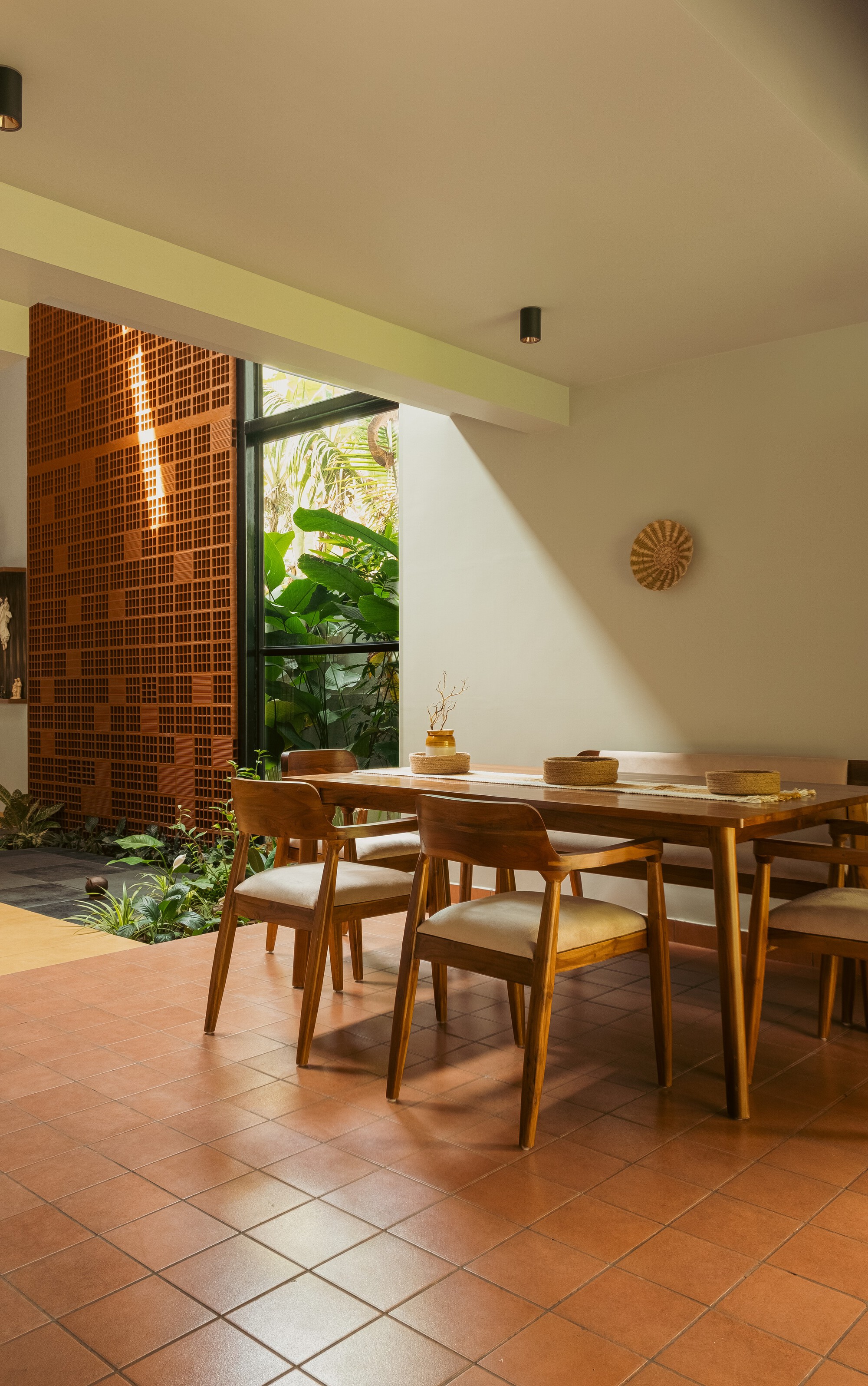 Timber dining table and chairs on terracotta tiles beside a perforated brick screen and a window of banana leaves, with afternoon sun on the wall