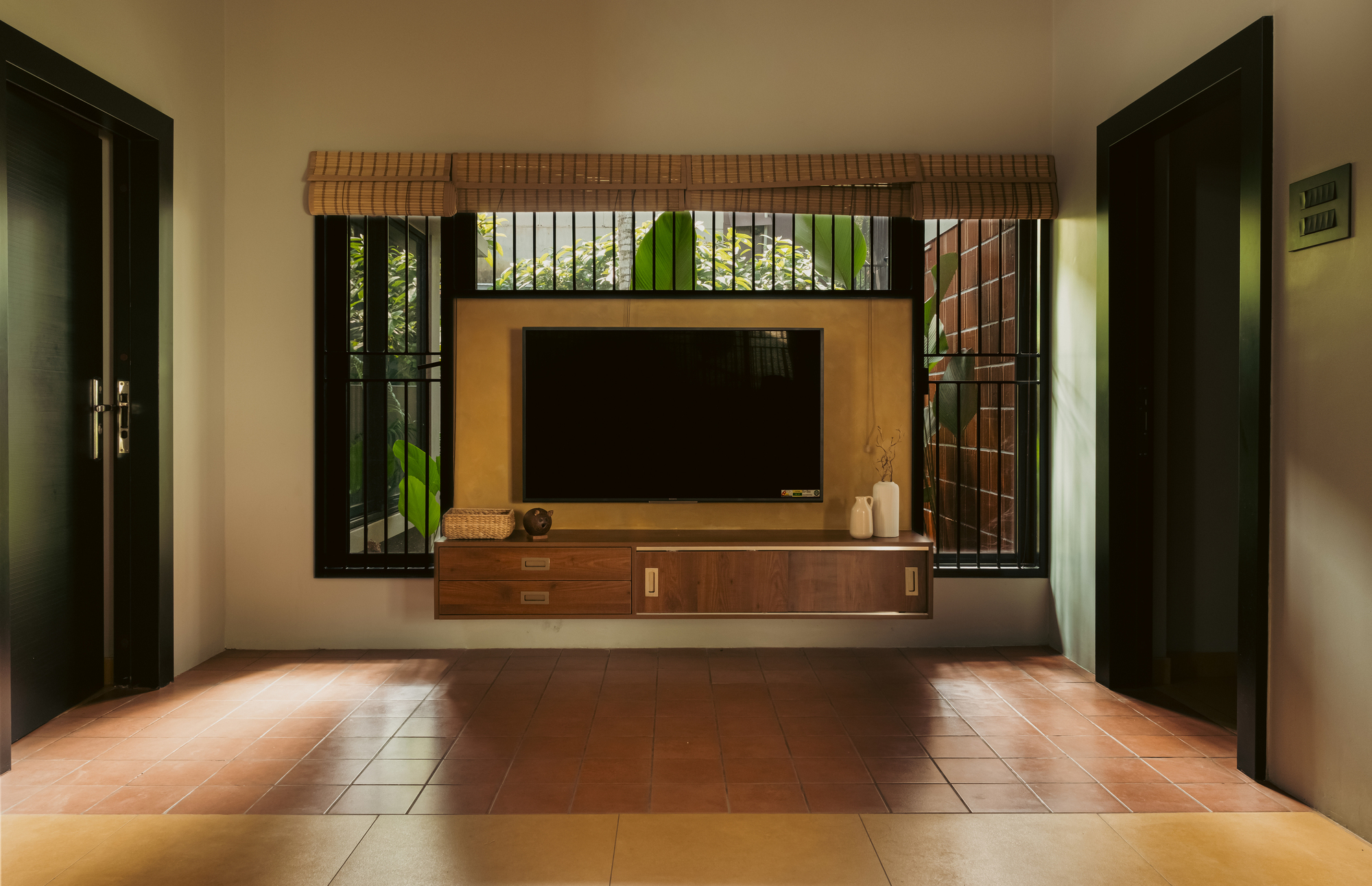 Wall-hung timber media console framed by barred windows and bamboo blind, with sunlight patches across terracotta floor tiles