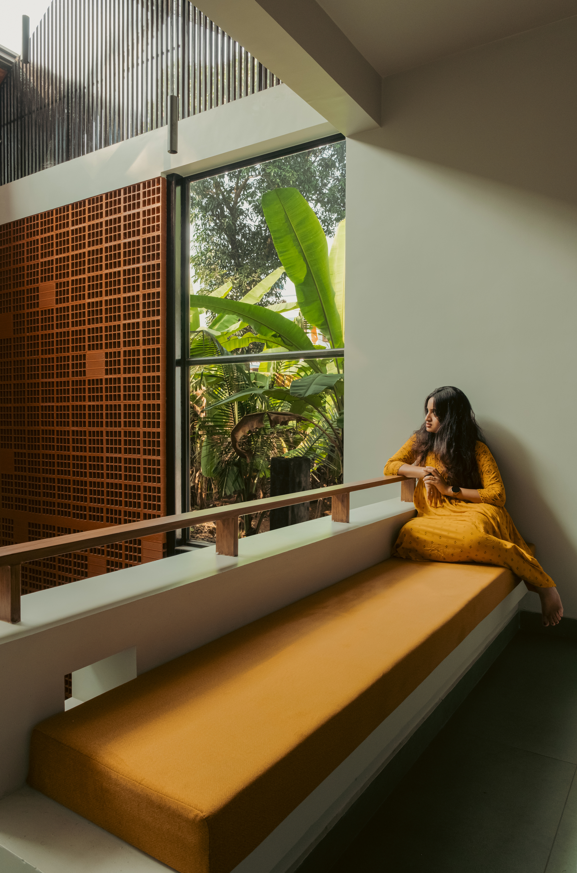 Woman in a yellow dress sits on a built-in ochre window seat beside a perforated brick screen and banana plants