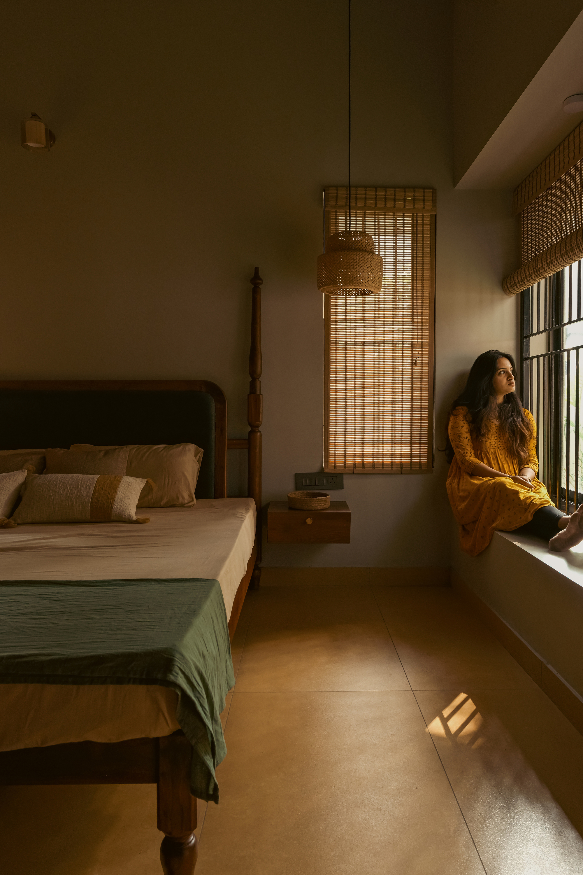 Bedroom with a four-poster timber bed, bamboo pendant lamp and woven blinds, a woman seated on the window ledge in soft light