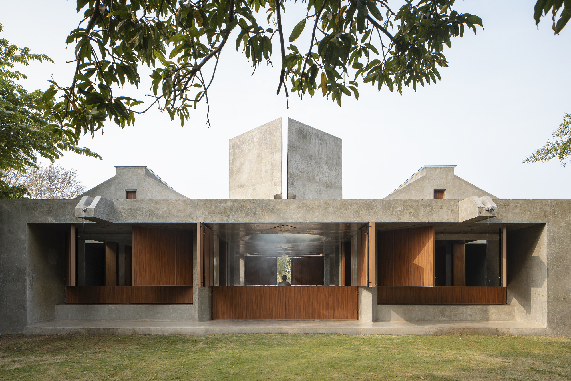 Front elevation of a low concrete pavilion with open timber shutters and a central concrete tower, framed by overhanging tree branches in hazy light