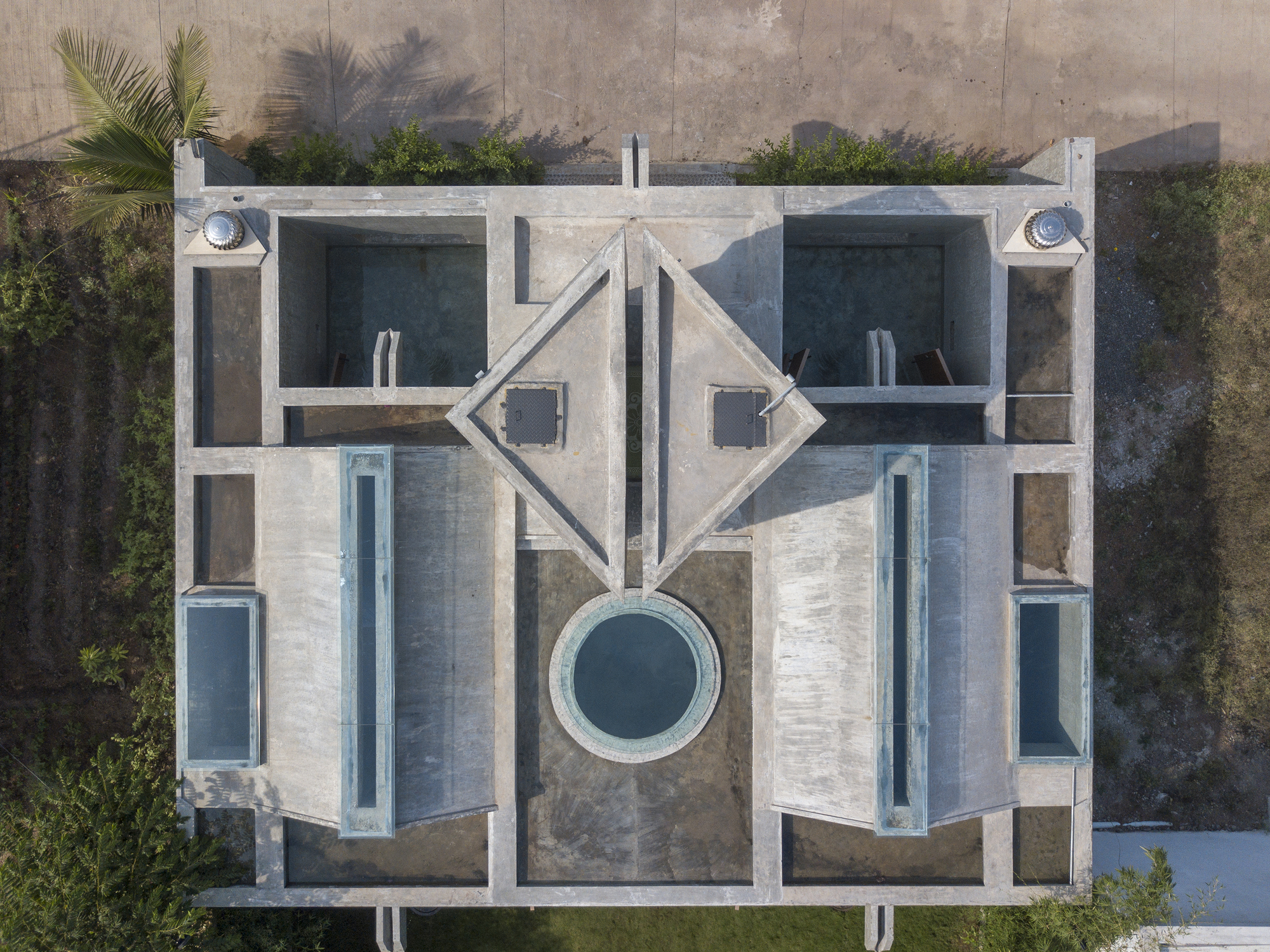 Top-down view of the concrete roof with triangular skylights, a circular pool and rooftop basins, seen with palm shadows at the edges