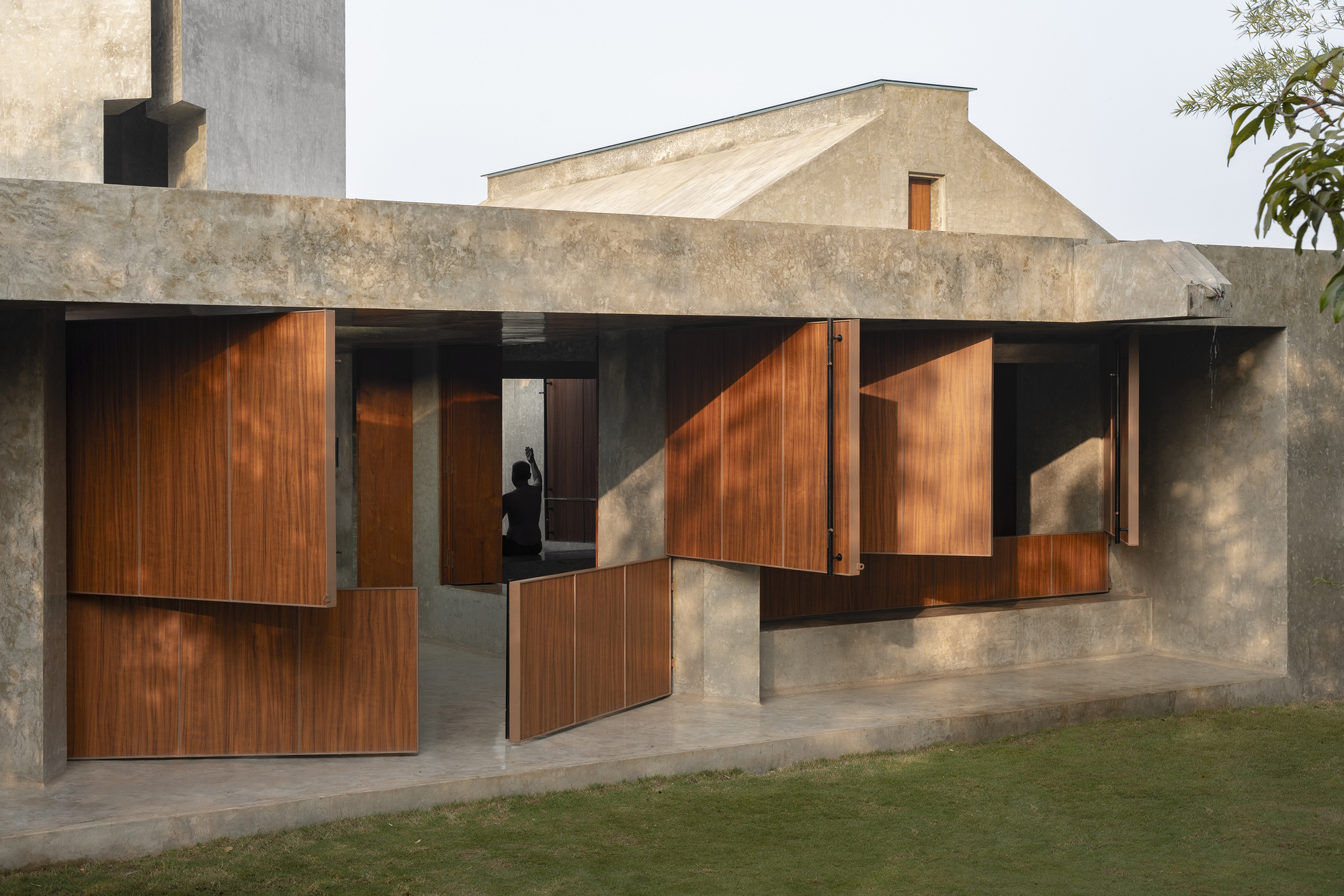 Corner of the concrete pavilion with pivoting timber shutters propped open and a figure seated inside, dappled tree shade on the lawn side