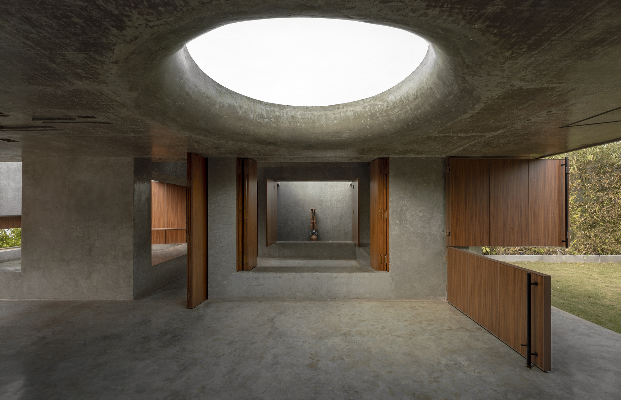 Interior of the concrete room with a round oculus overhead, pivoting timber doors and a figure doing a handstand in the far alcove