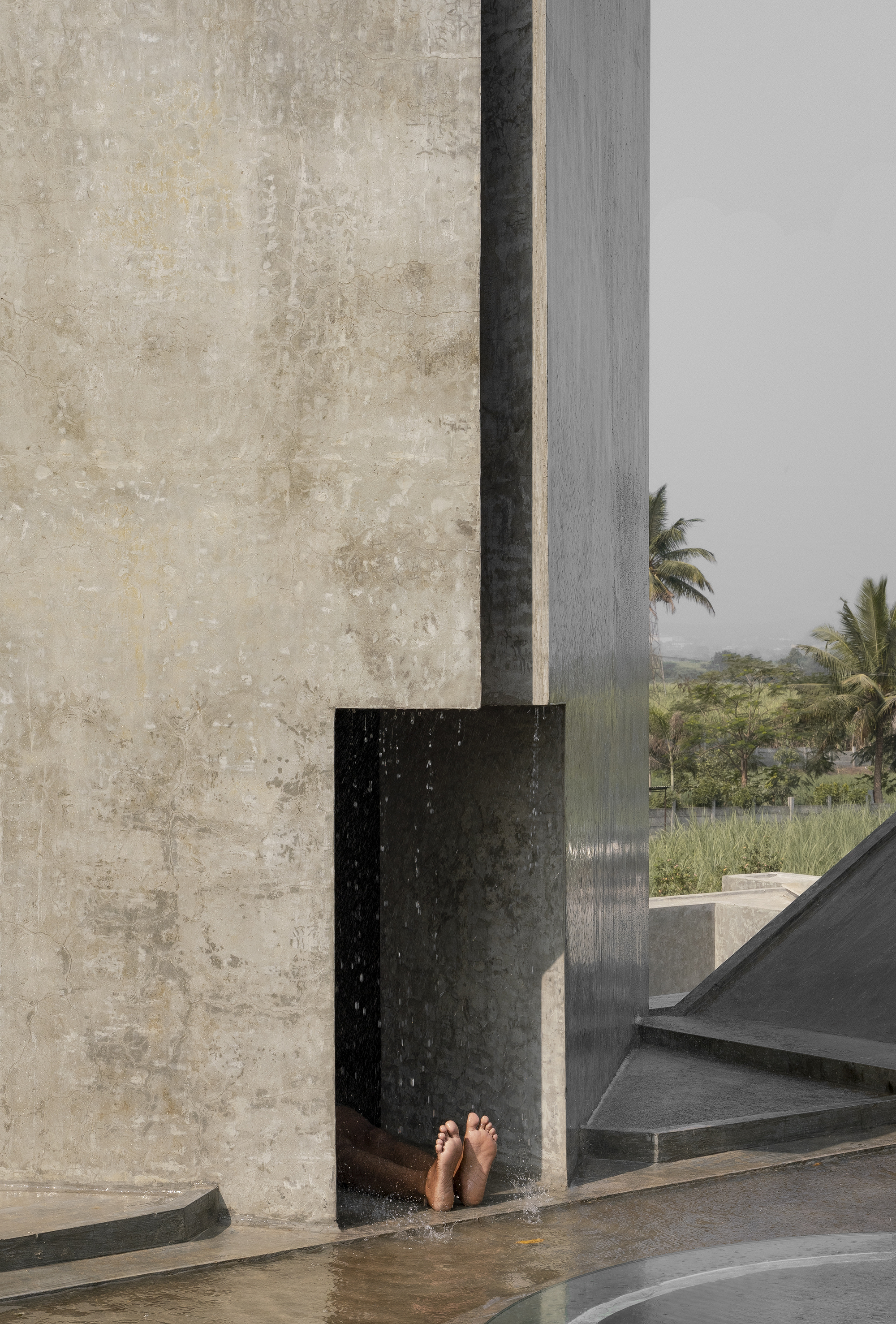 Concrete wall with a recessed niche where water falls over a person's bare feet, with palms and a pitched roof edge in hazy air