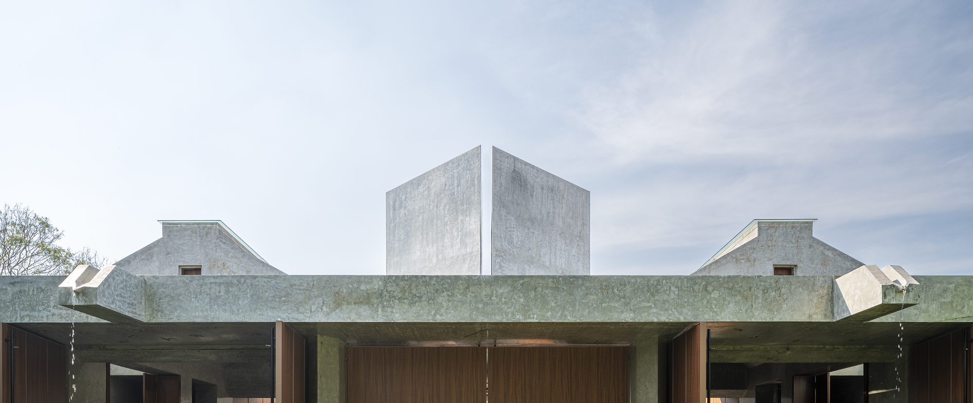 Concrete roofline with two small gabled dormers and a split angular volume rising above timber doors under a pale hazy sky