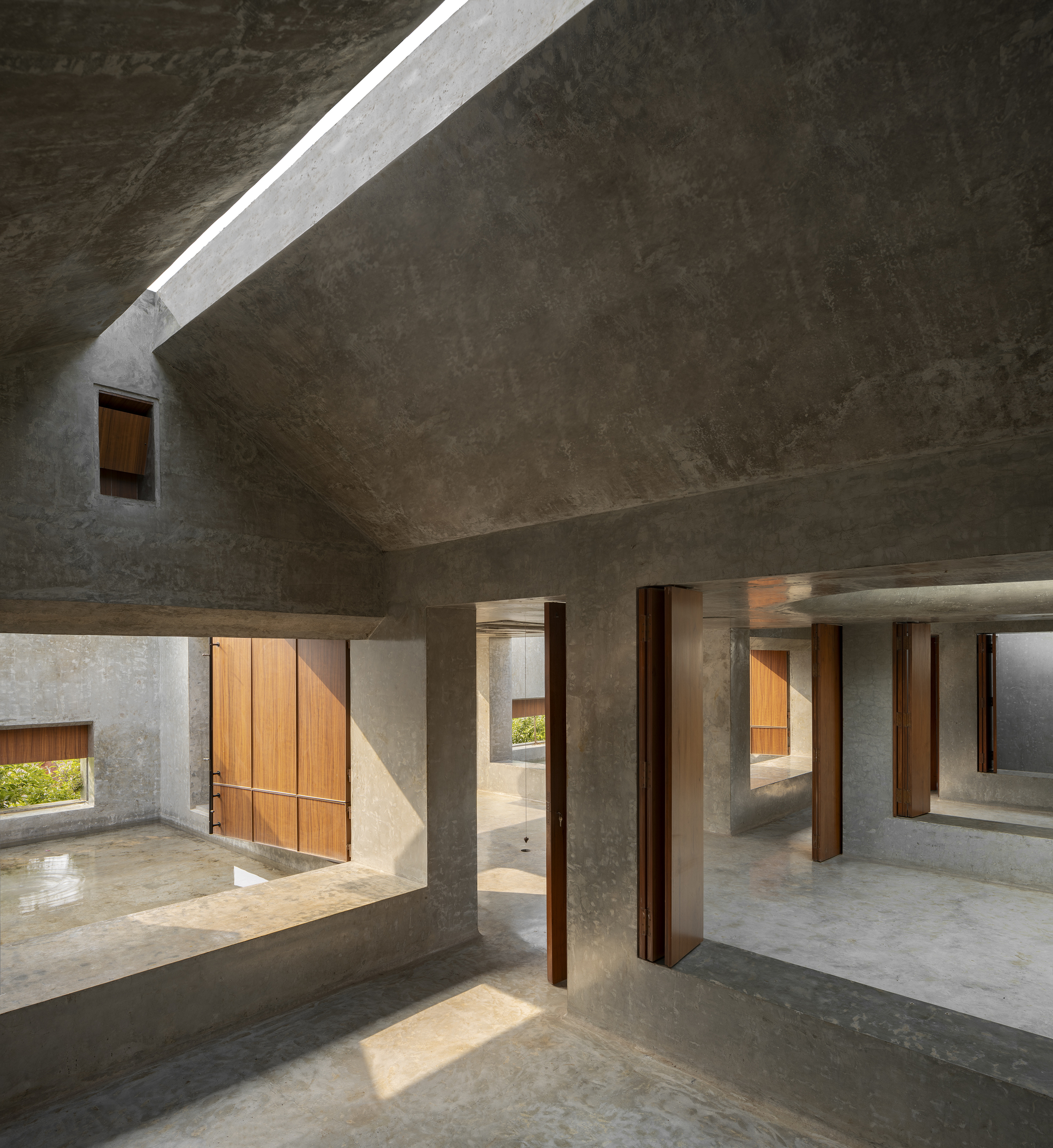 Interior of rough concrete rooms with a pitched ceiling slit of daylight and tall timber shutters open to sunlit floors