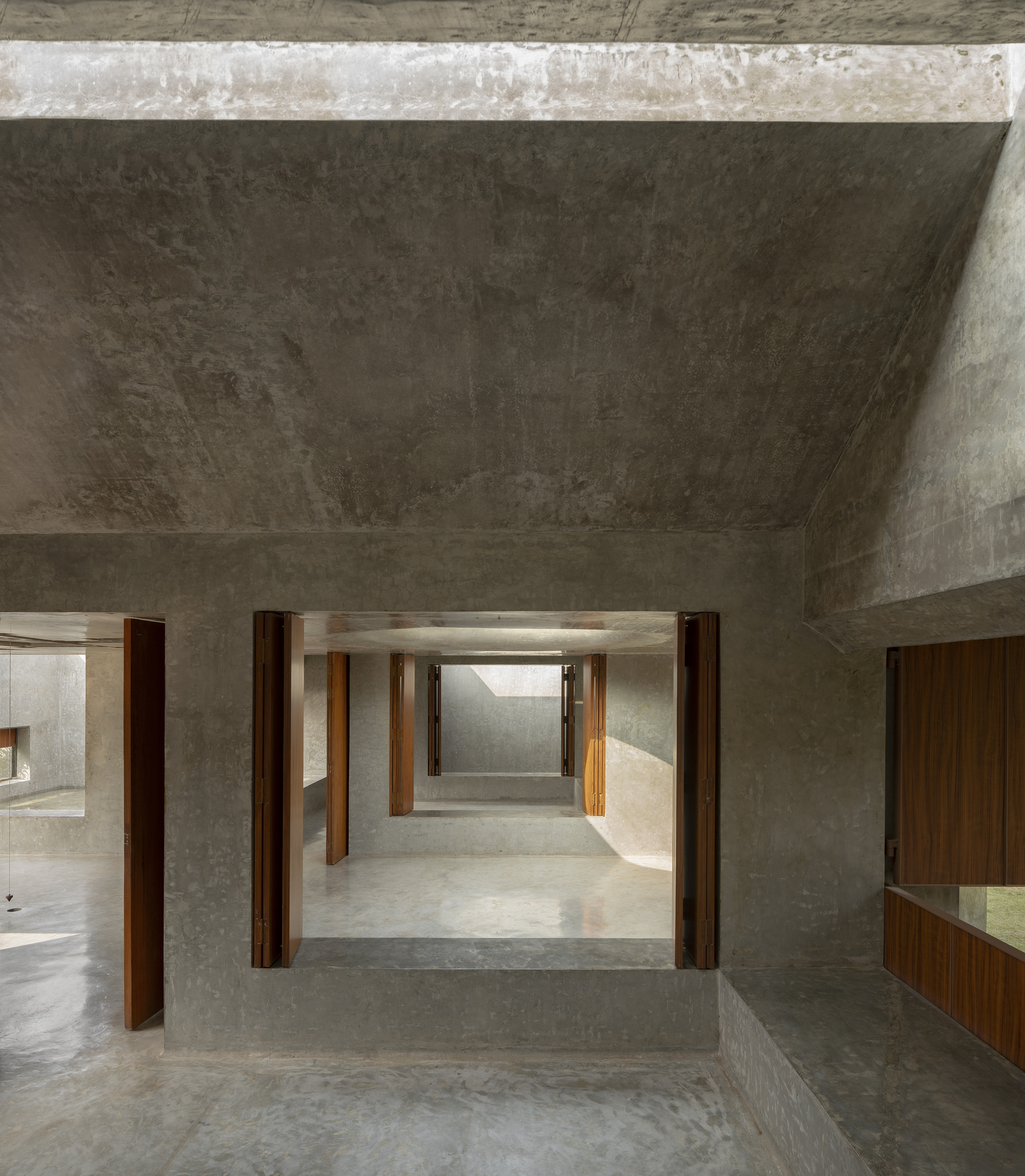 Enfilade of concrete rooms seen through a square opening framed by open timber shutters, with soft light on the polished floor