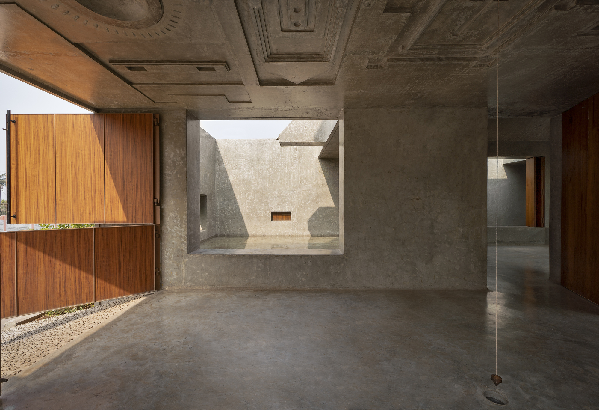 Concrete room with a coffered ceiling, a square window onto a shaded courtyard and a lifted timber shutter letting in afternoon sun