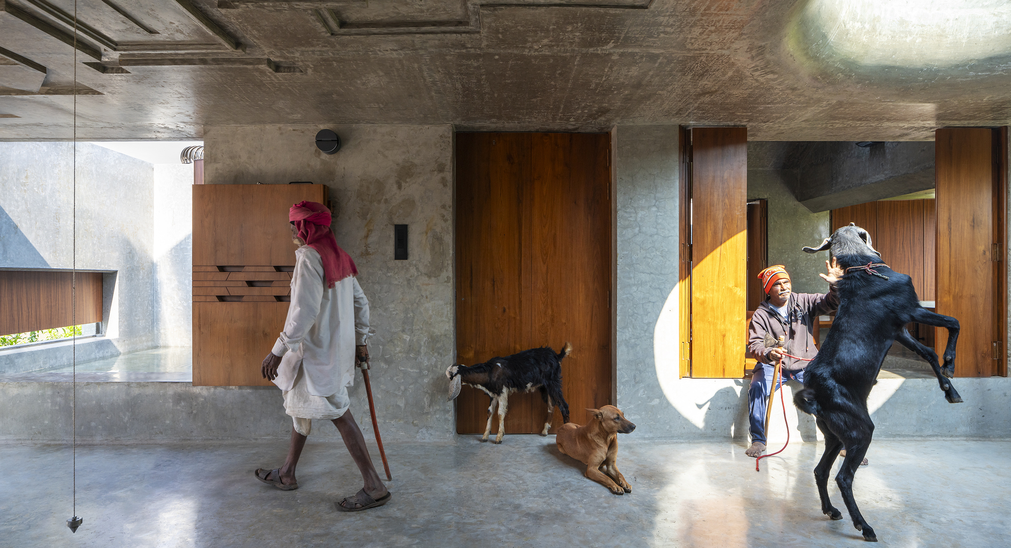 Two herders with goats and a resting dog on a polished concrete floor in front of timber doors in daylight