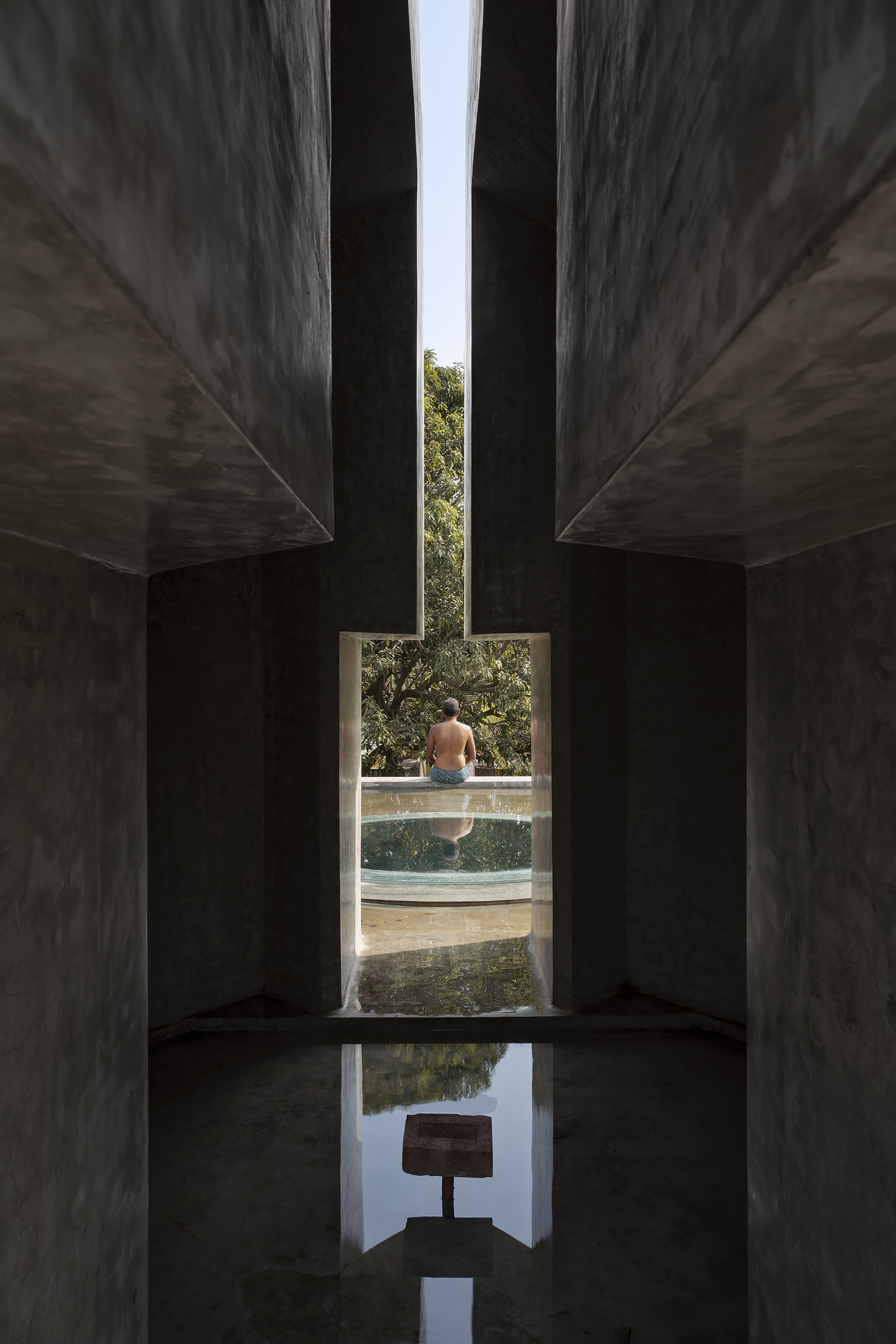 Narrow concrete passage framing a seated figure beside a round pool, with a leafy tree beyond and a reflecting water channel below