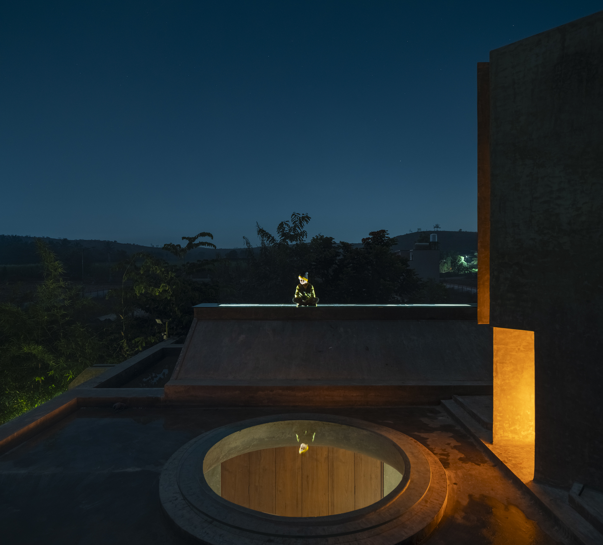 Rooftop at dusk with a circular skylight rimmed in stone, a crouching figure on the parapet and a lit slot in the concrete wall