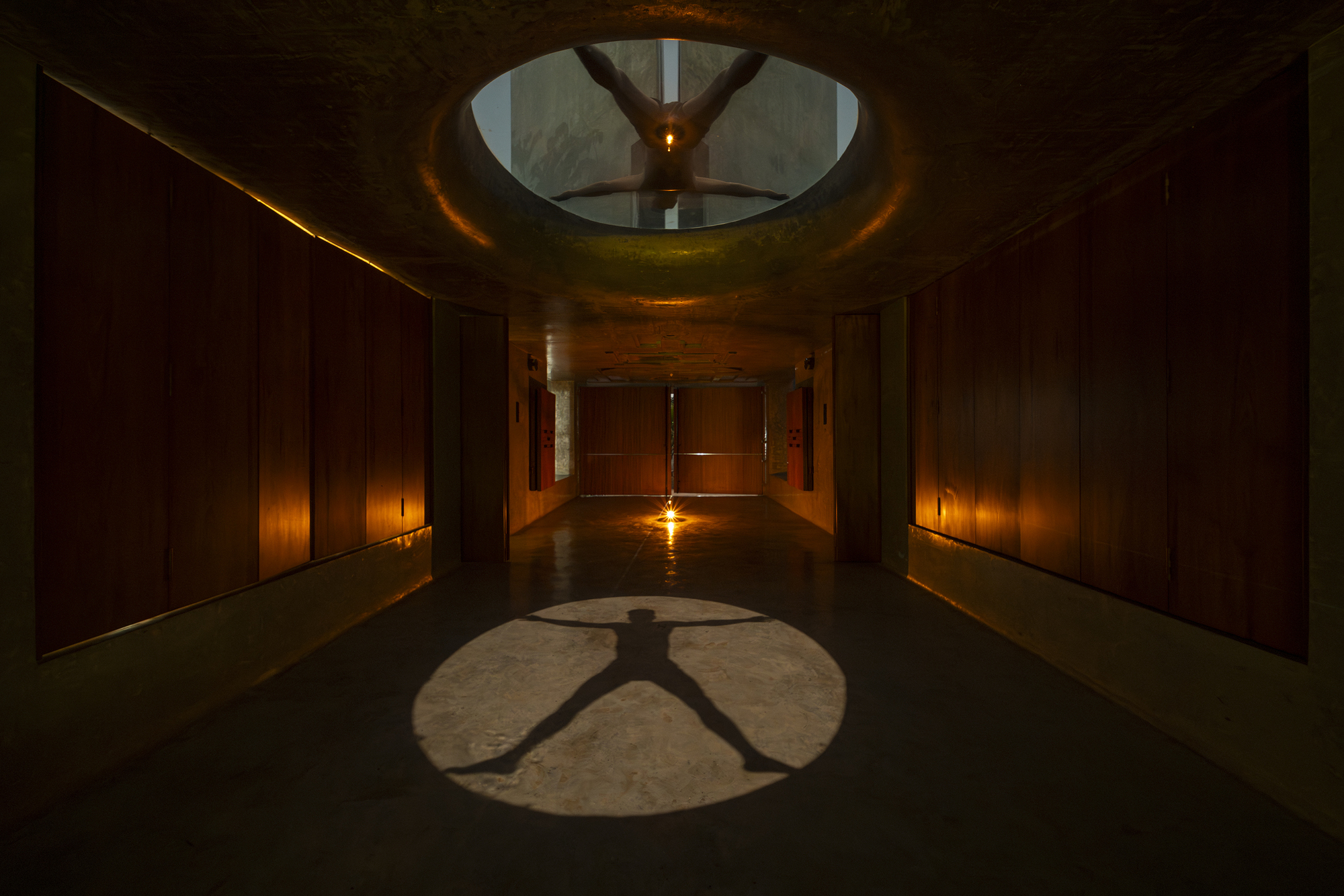 Corridor lined with timber panels, where a circular skylight casts a spread-armed figure's shadow onto the polished floor in low light