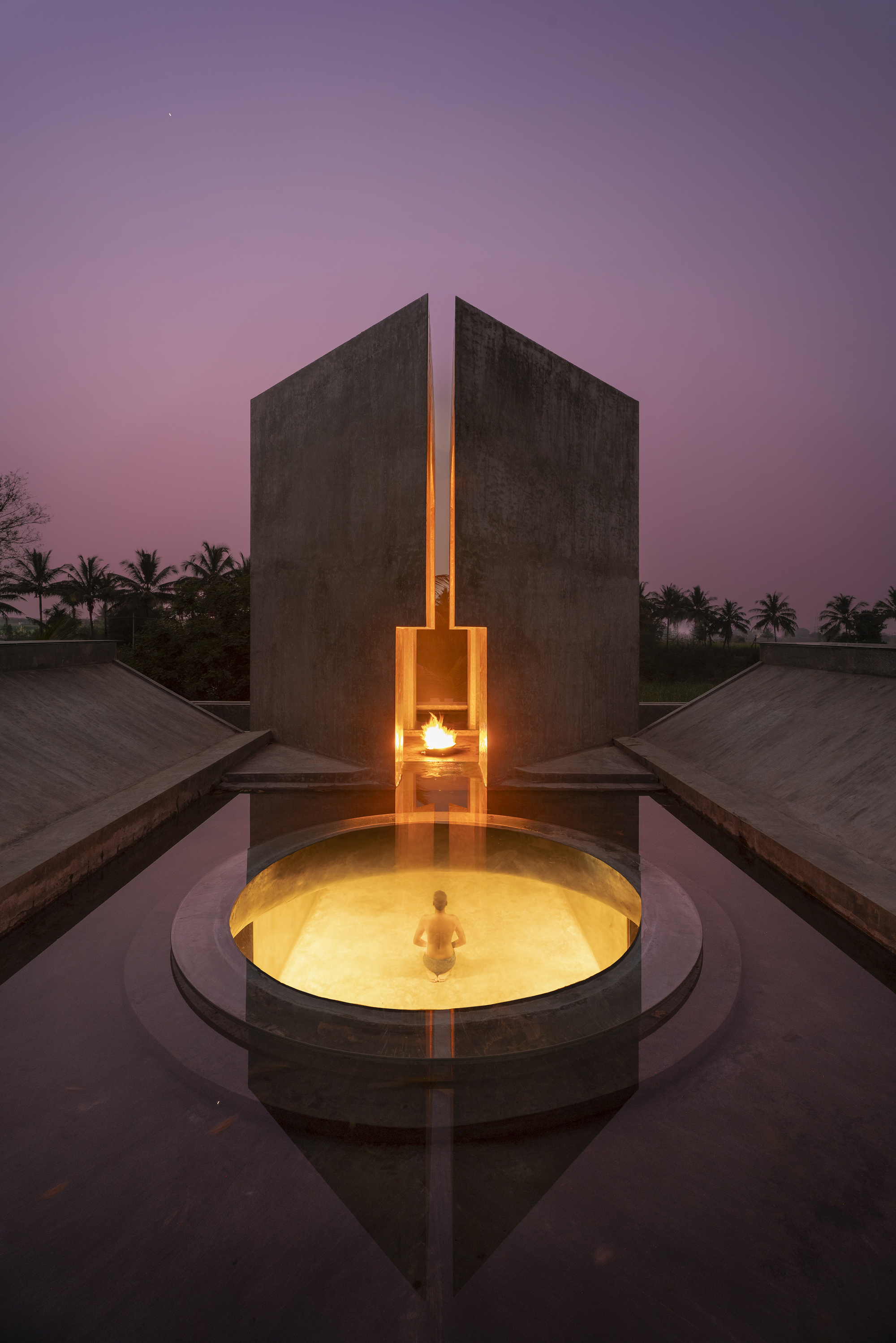 Twin concrete walls split by a glowing slot and flame, above a circular pool with a bather at purple dusk