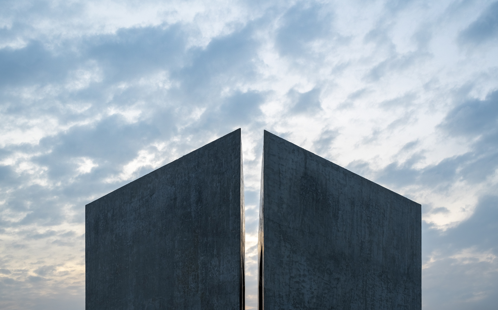 Two angled concrete slabs separated by a thin gap, rising against a sky of scattered evening clouds