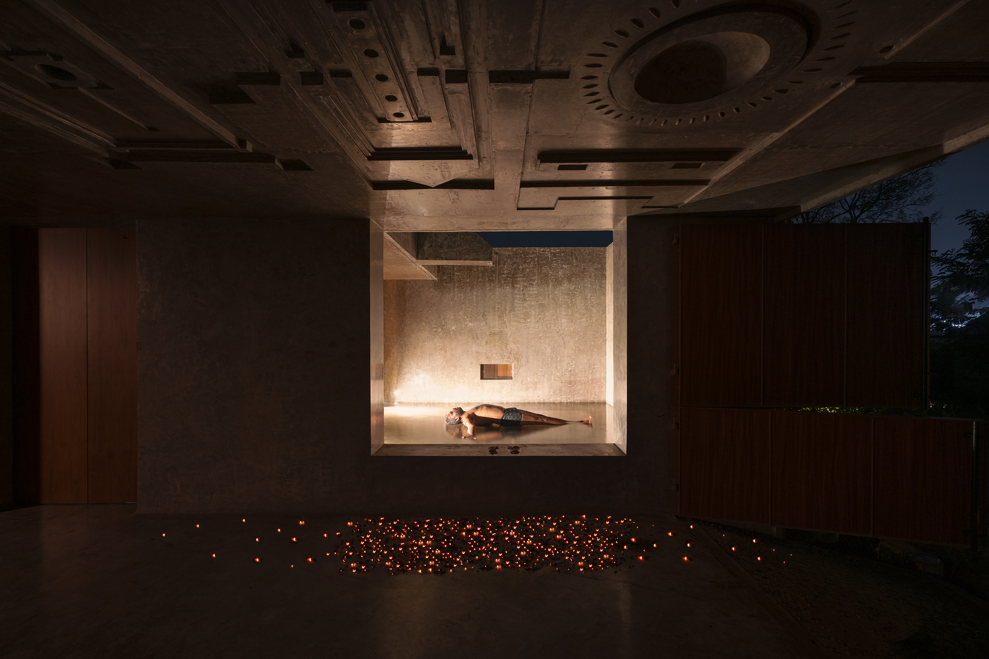 Recessed concrete niche with a reclining figure, a carved ceiling above and scattered small lamps glowing on the floor at night