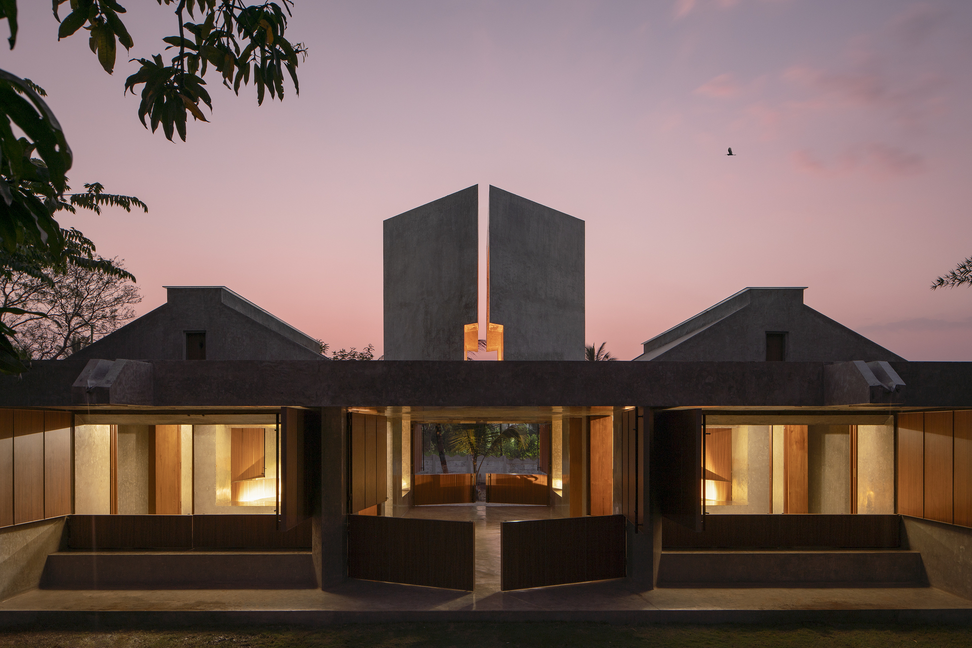 Symmetrical concrete pavilion with split central tower and open timber doors, seen at dusk under a pink sky with a bird overhead