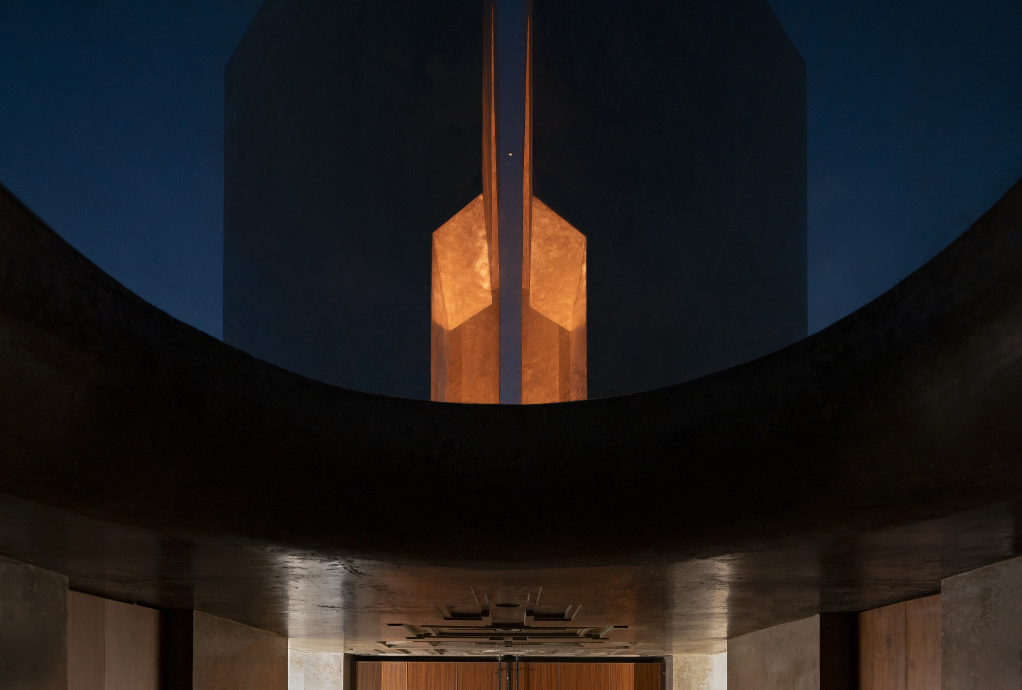 Looking up through a curved concrete opening at the split tower, its glowing amber interior lit against the night sky