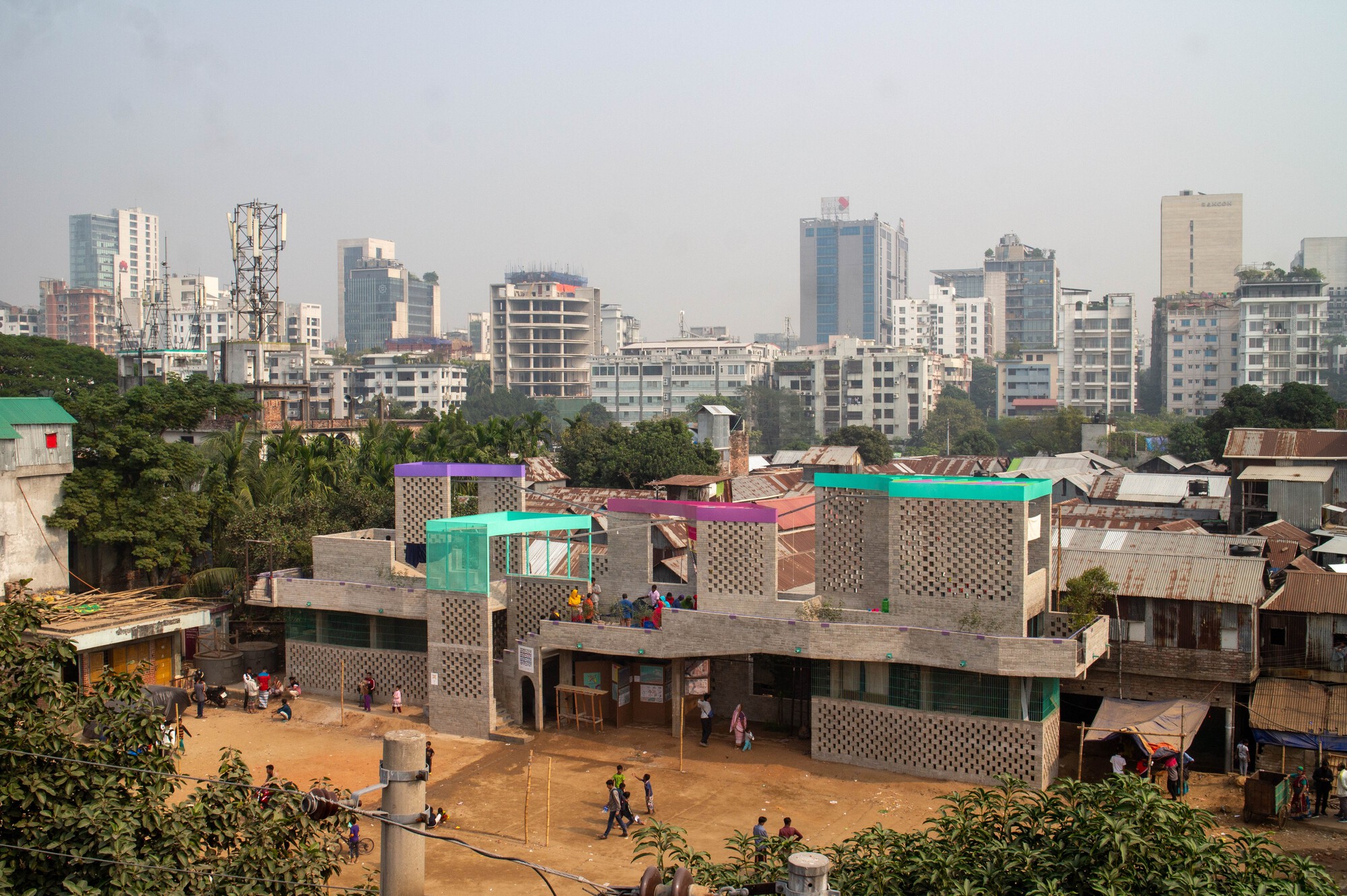 Elevated view of the perforated brick building with teal, purple and pink roof canopies, surrounded by tin-roofed houses and a dusty open ground in hazy dayligh