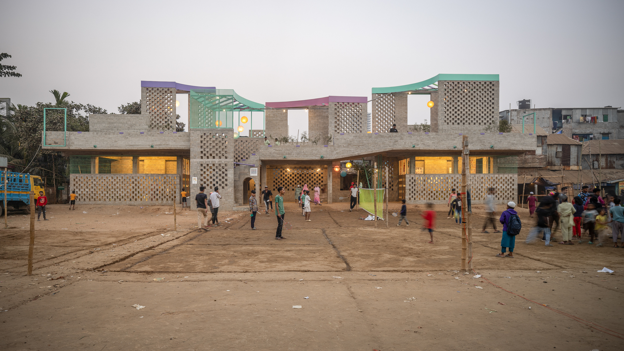 Front view of the long brick building with lattice walls and colored roof frames as children play on the bare ground at dusk